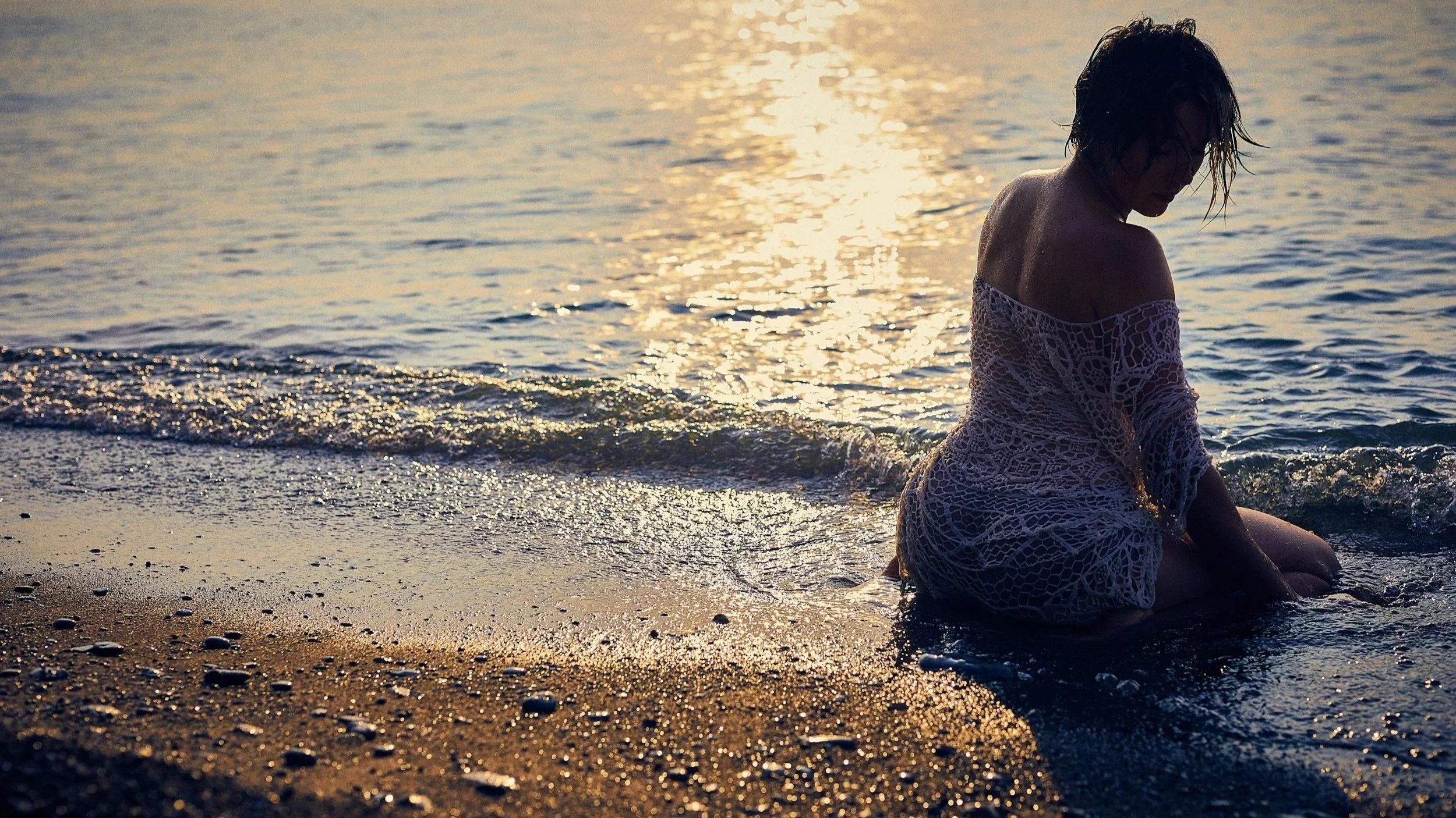 A woman in a white lace dress sitting on a beach at sunset, facing the water with her head bowed and wet hair, with calm waves and golden sunlight reflecting on the water.