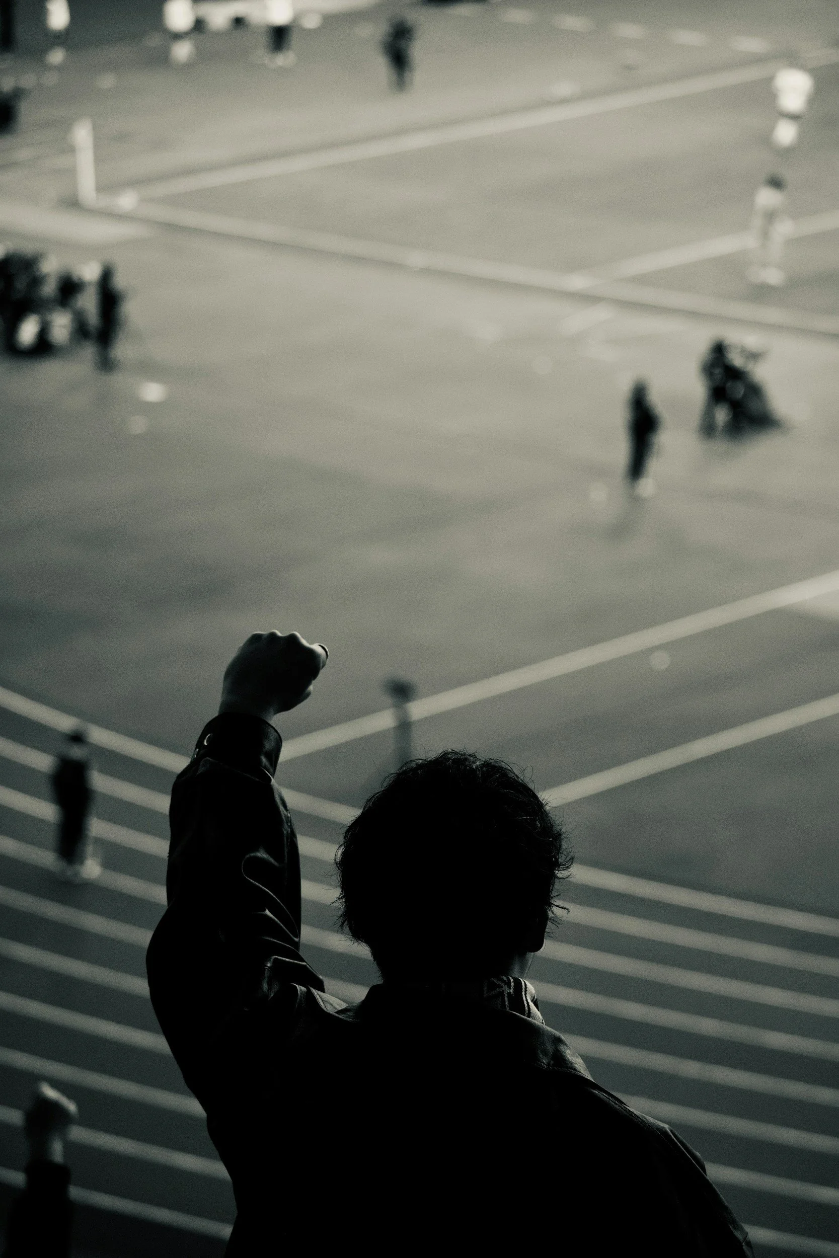 A person with curly hair and wearing a leather jacket is raising their fist while overlooking a sports field with blurred figures playing or walking in the background.