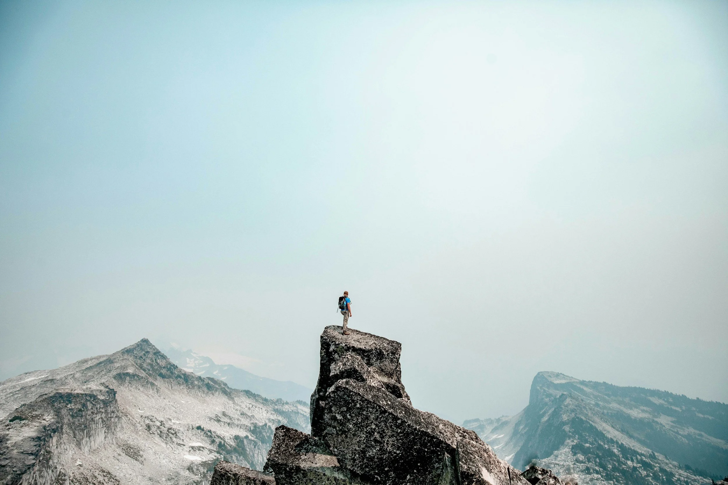 A lone hiker standing on a large rock formation in a mountain range with snow-capped peaks and a cloudy sky in the background.