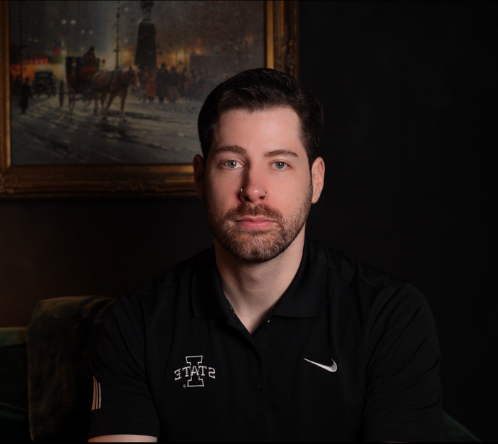 A young man with dark hair and a beard, wearing a black polo shirt with a Nike logo and Indiana State University logo, sitting in a dimly lit room with a framed painting of horses and a city scene hanging on the wall behind him.