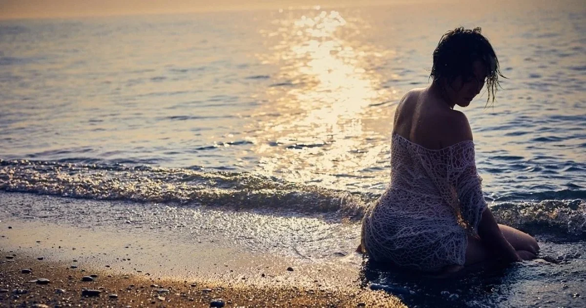 A woman sitting on the beach in shallow water during sunset, wearing a lace dress with wet hair, facing sideways with her head bowed.