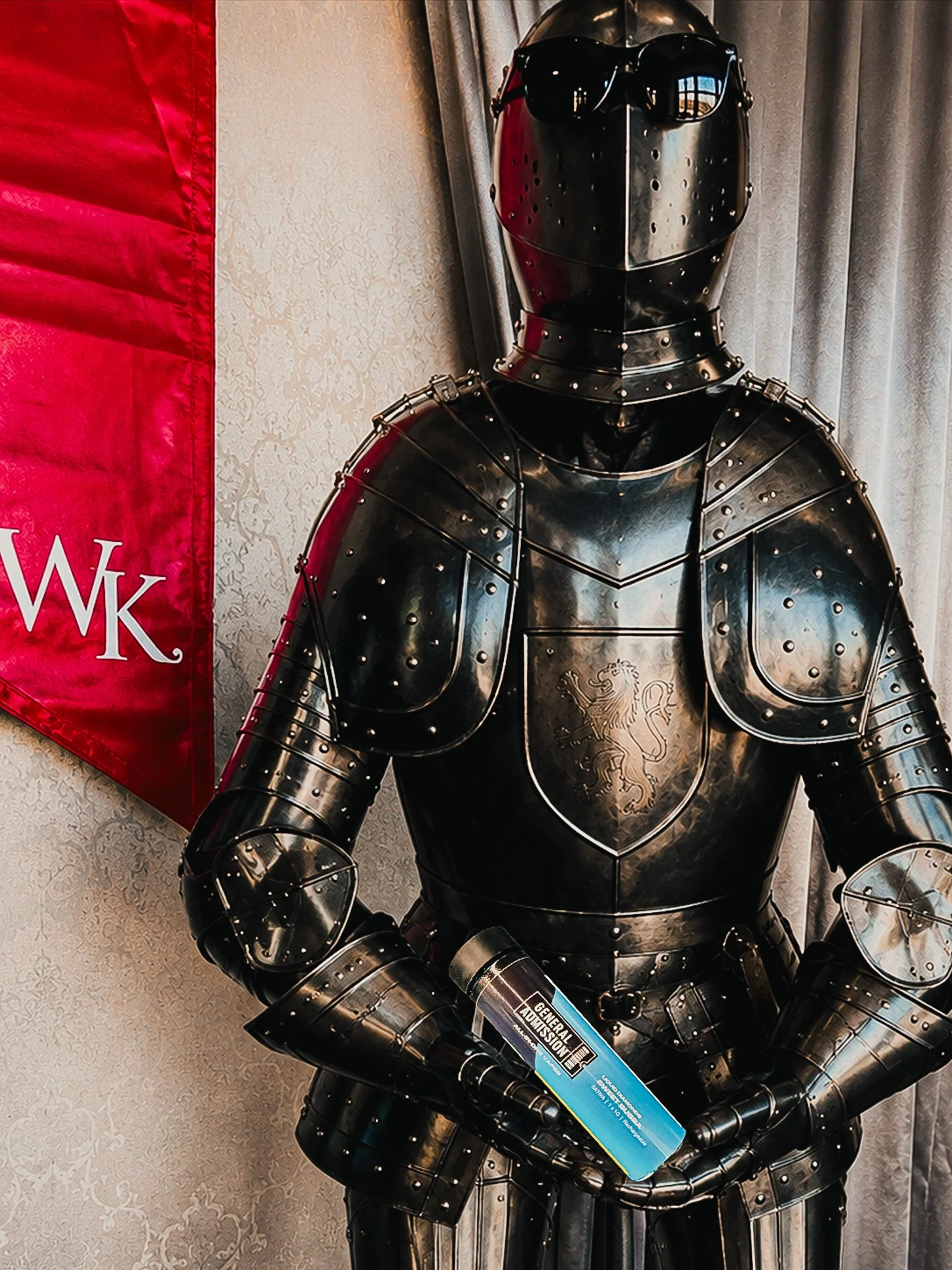 A suit of black armor with a lion crest emblem on the chest, displayed indoors beside a red banner with the letters W and K, and a pair of black sunglasses on the helmet.