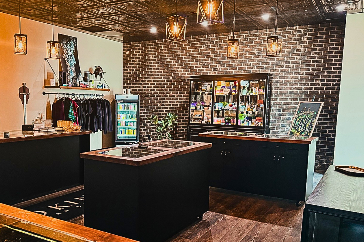 Interior of a retail store with brick walls, hanging pendant lights, a clothing rack with jackets, a beverage refrigerator, a display case with various products, and a framed sign that reads "Sylvan Lake"