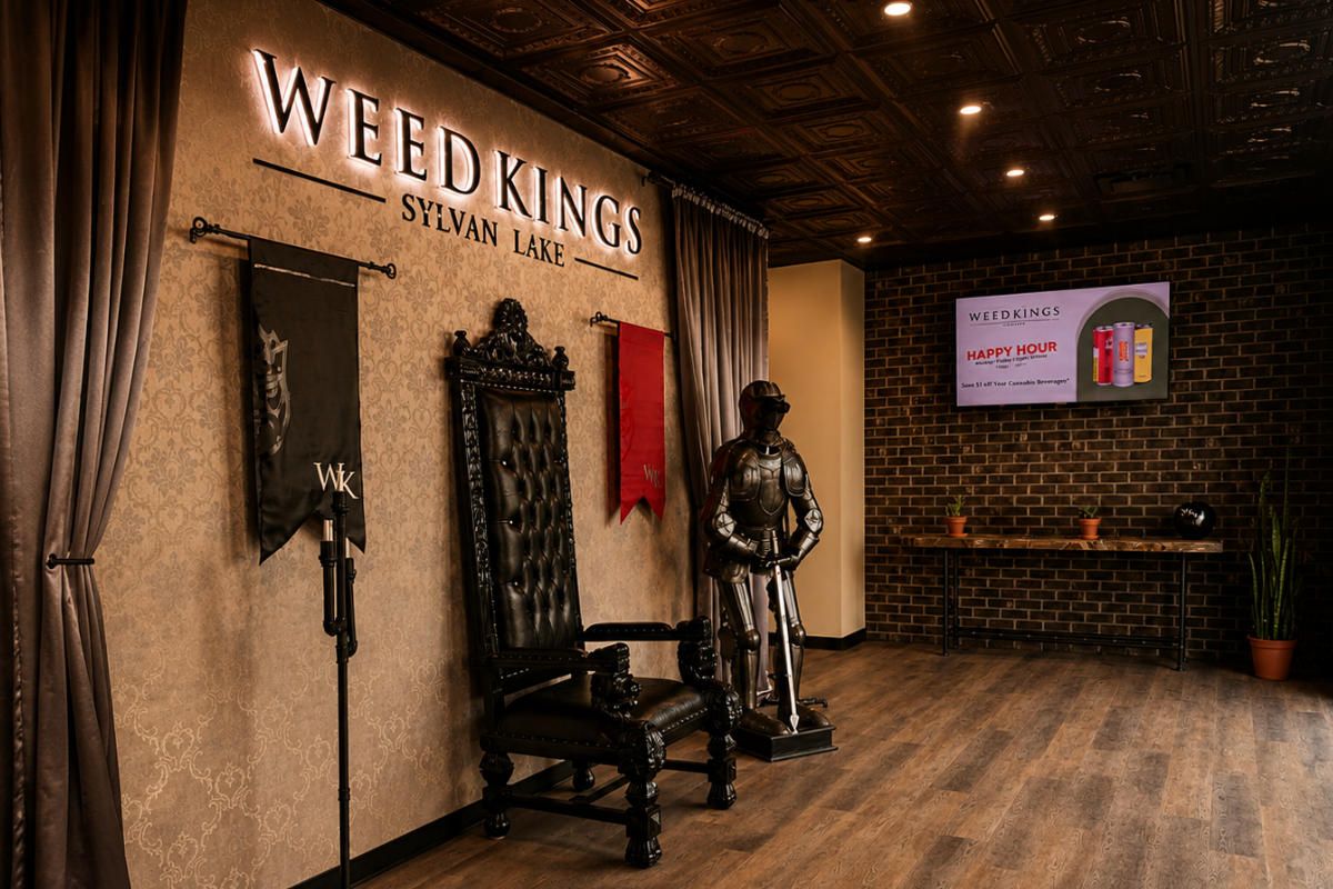 Interior of a venue with a large sign that reads "WEED KINGS SYLVAN LAKE." There are curtains on the left, a tall ornate black throne-style chair, a mannequin dressed as a robot or armored figure, a hanging flag, and a digital display on a brick wall advertising happy hour drinks with colorful cans.