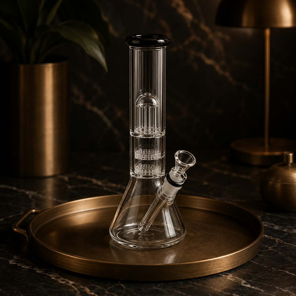A glass water pipe, or bong, with a straight tube design and percolator chambers, placed on a round brass tray on a dark marble surface with warm lighting and a potted plant in the background.