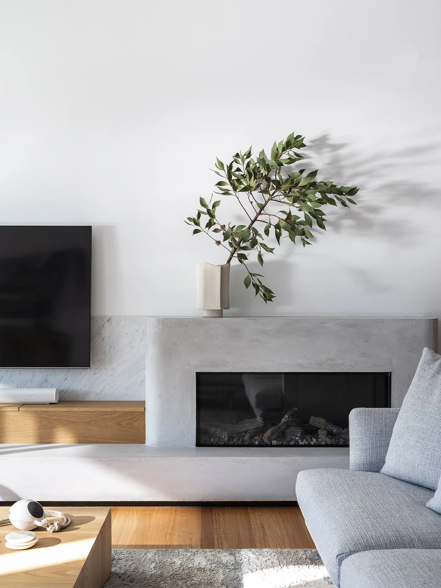 Modern living room with a minimalist design featuring a concrete fireplace, a flat-screen TV, a wooden coffee table with decorative objects, and a light gray sofa. A vase with a green leafy branch is placed on the fireplace mantel, casting a shadow on the white wall.