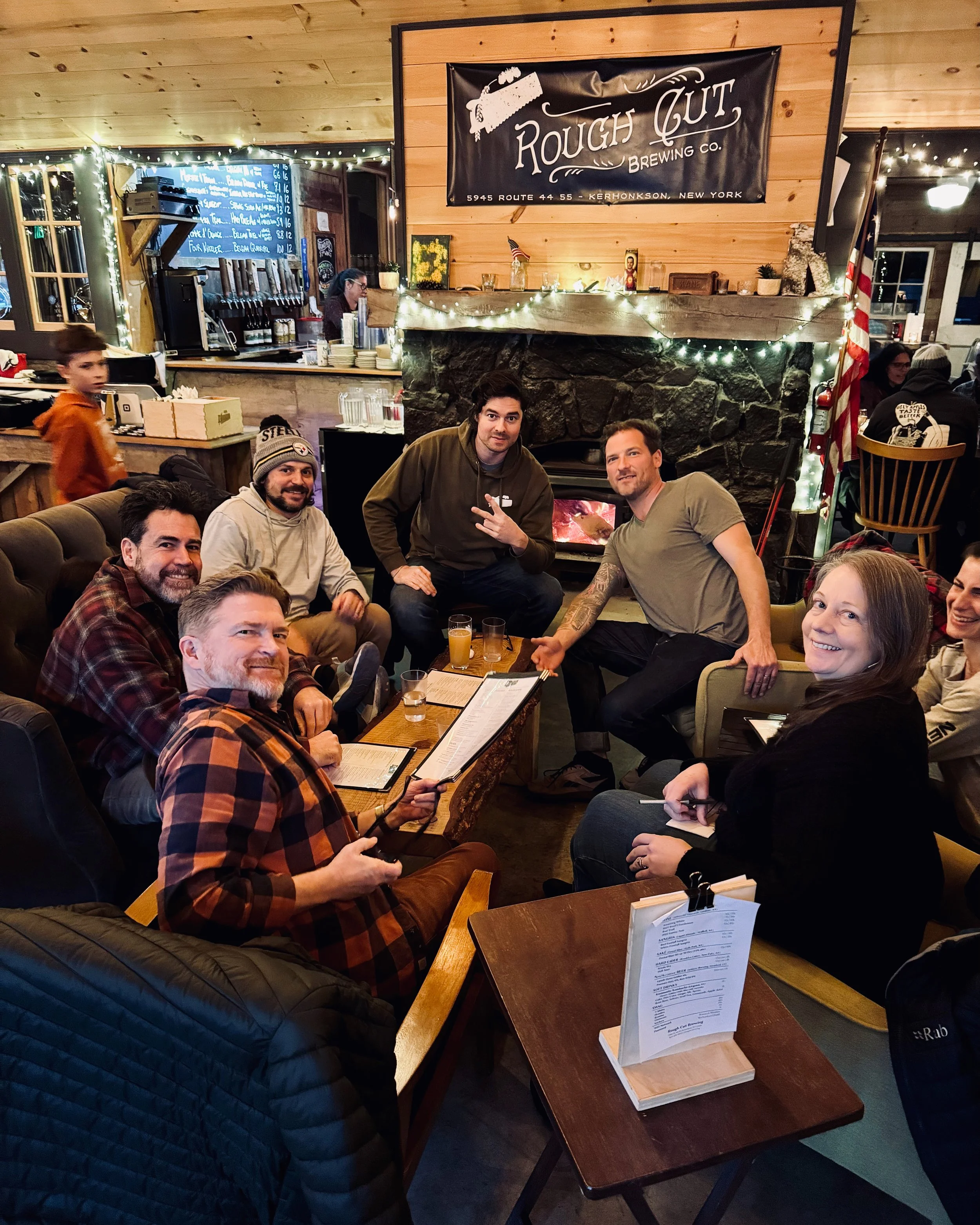 A group of eight people sitting around a coffee table in a cozy, festive restaurant with wood-paneled walls, a stone fireplace decorated with string lights, and a banner reading "Rough Cut Brewing Co."