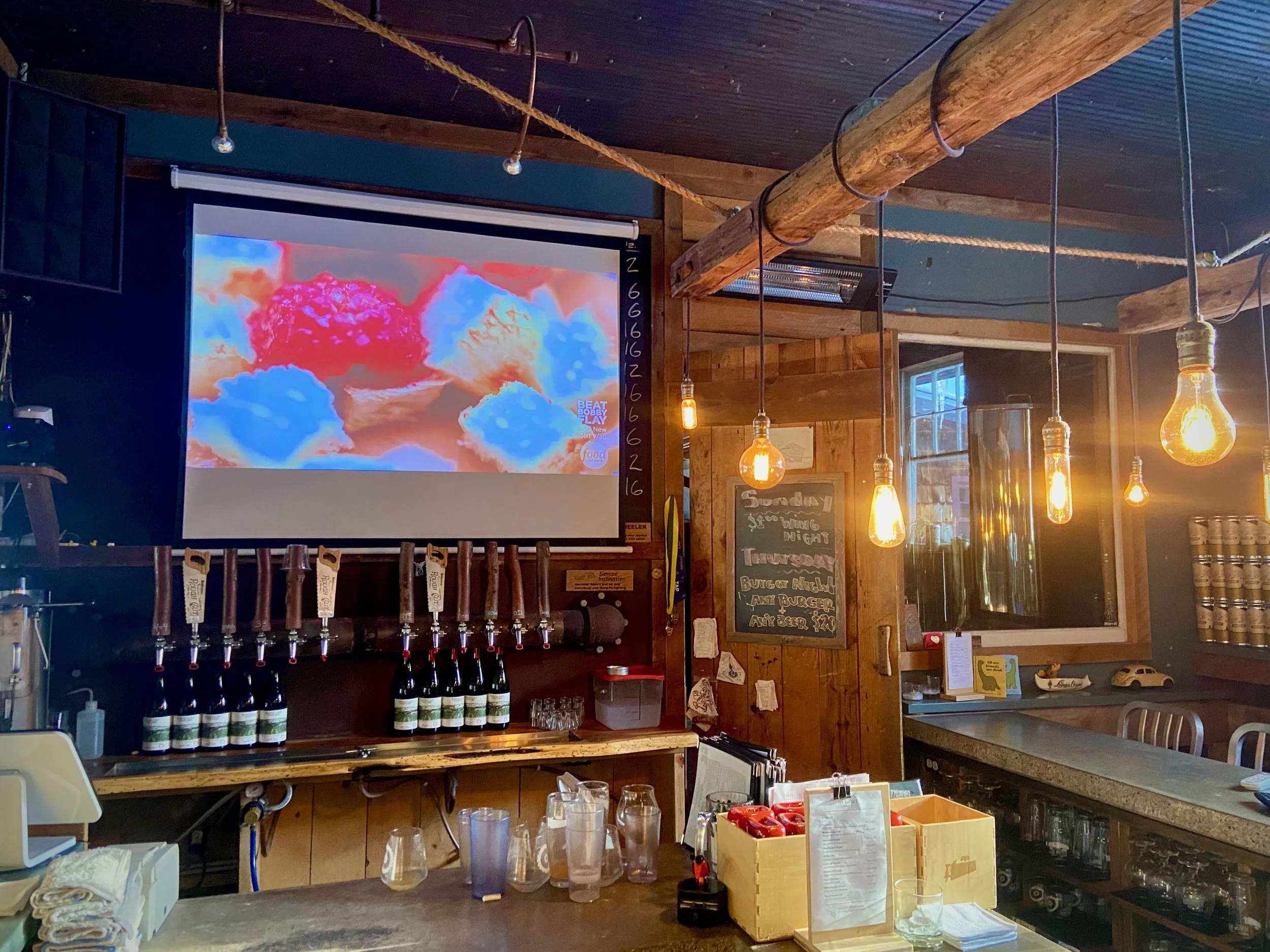 Inside a cozy restaurant with hanging Edison bulbs, a large TV screen showing colorful fluffy clouds with a red cloud in the center, and beer taps below the screen. The wooden walls and furniture create a warm atmosphere, with a small chalkboard menu and various items on the counter.