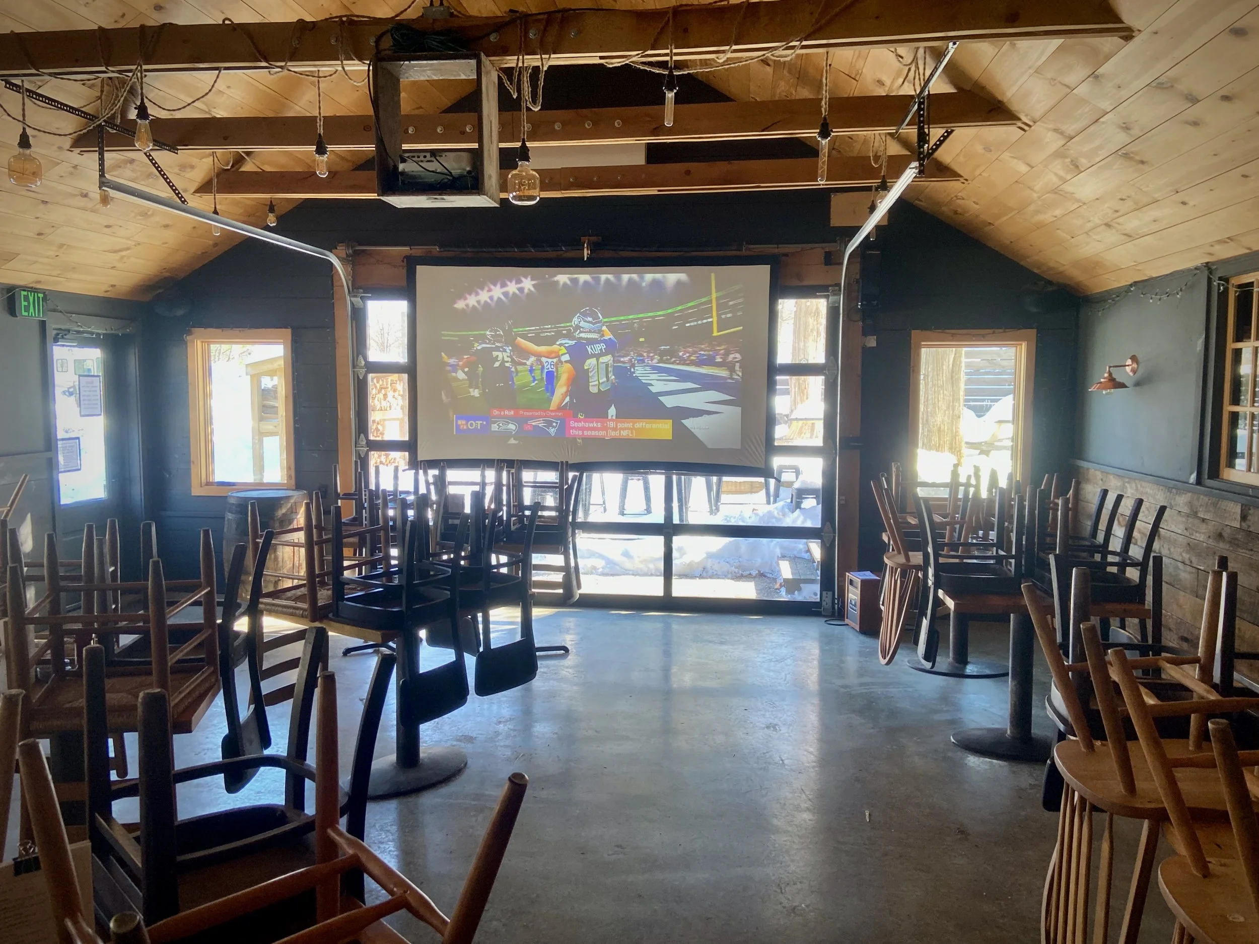 An empty bar or restaurant with a large flat-screen TV showing a football game on the wall, with chairs stacked upside down on tables, and sunlight coming through windows testing out the sports projector screen before opening