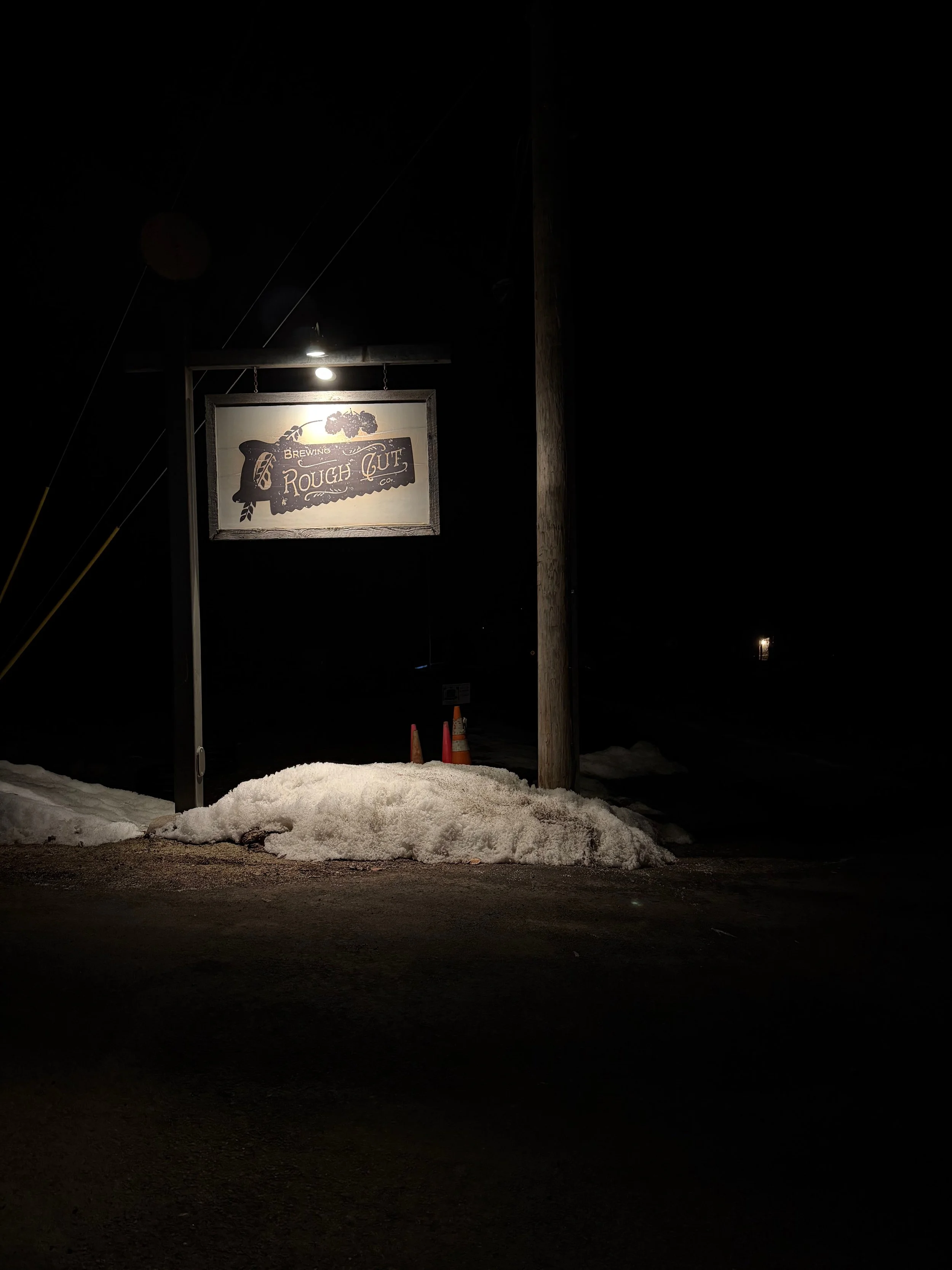 Nighttime photo of a lit sign for 'Rough Cut Brewing Co.' with snow and ice on the ground, and traffic cones in the background.