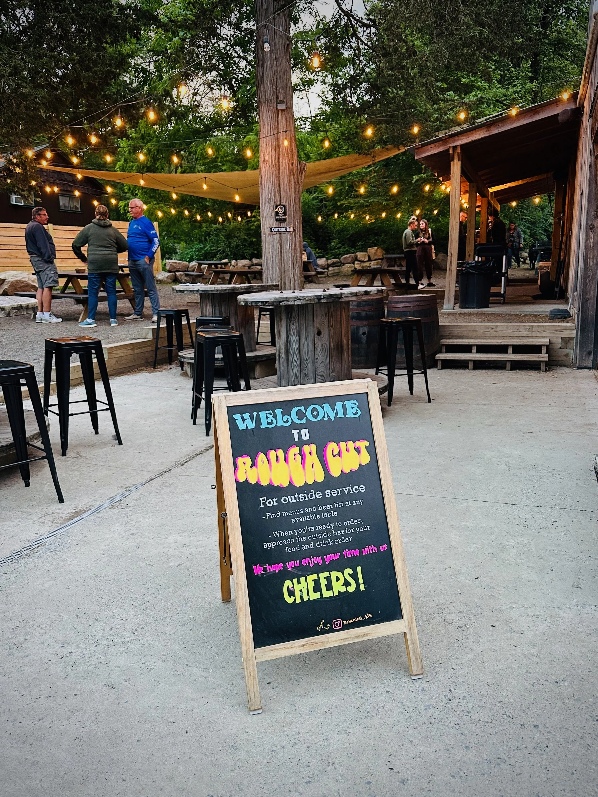 Outdoor restaurant area with string lights, wooden tables, and a chalkboard sign welcoming guests to Rough Cut. The sign mentions outside service, menu and beer list availability, and a message inviting guests to enjoy their time. There are a few groups of people socializing in the background.