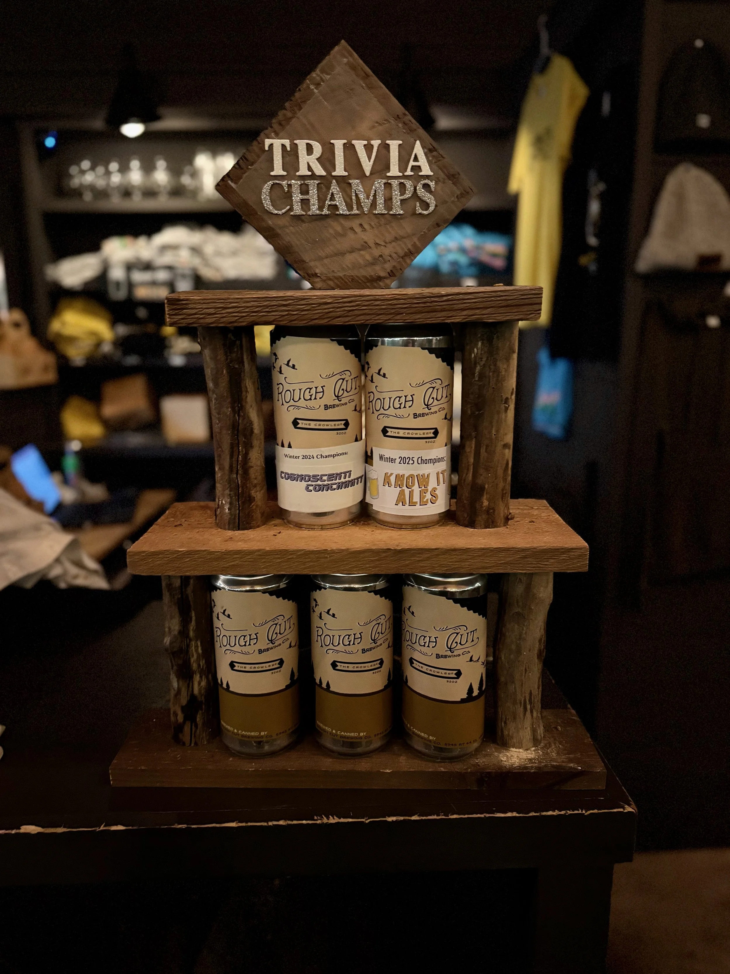 A wooden display rack with cans of Rough Gulp brewing company beer, with a 'Trivia Champs' sign on top.