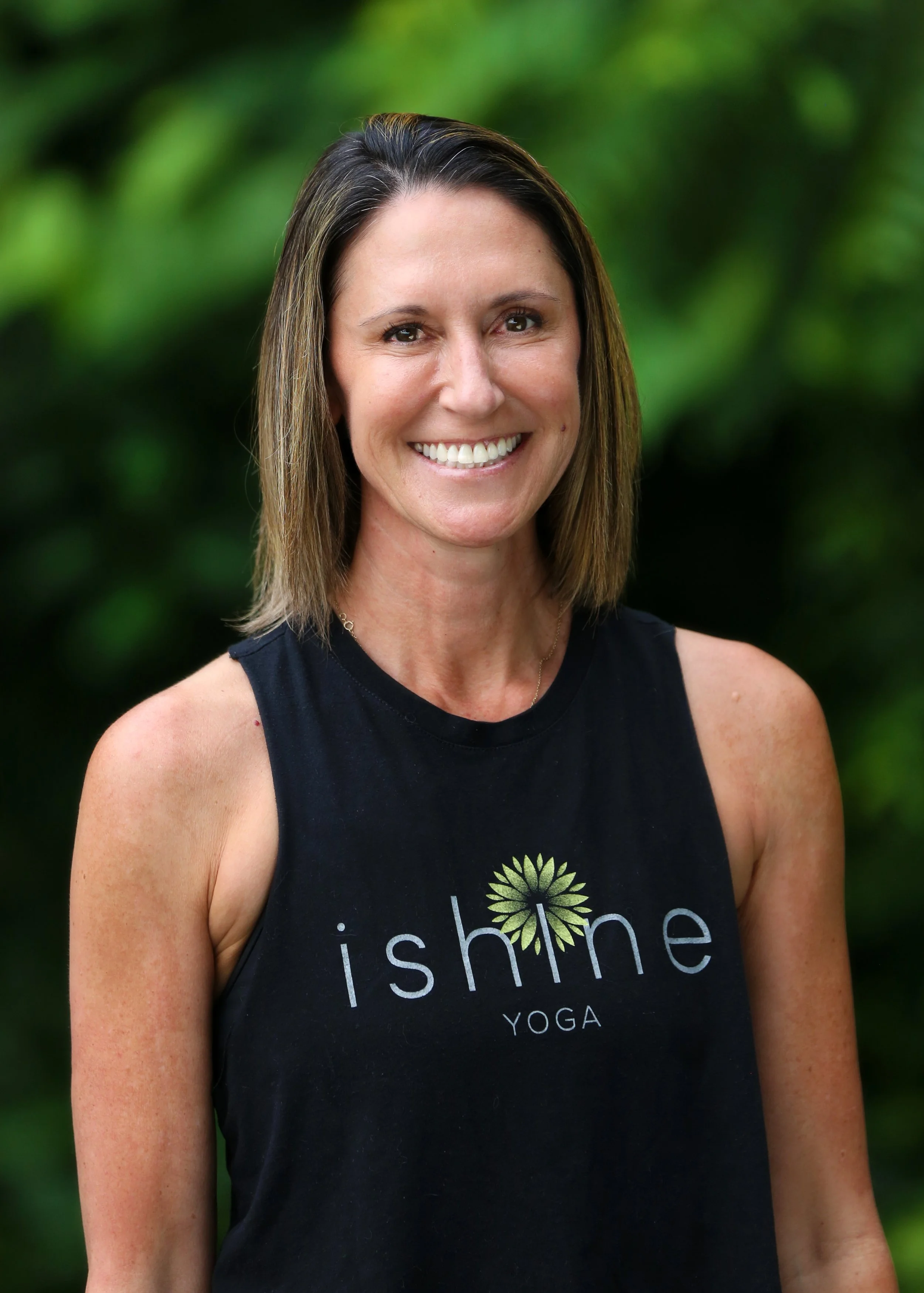 A smiling woman with shoulder-length brown hair wearing a black sleeveless shirt with the words 'ishine YOGA' and a dandelion logo, standing outdoors with a blurred green background.