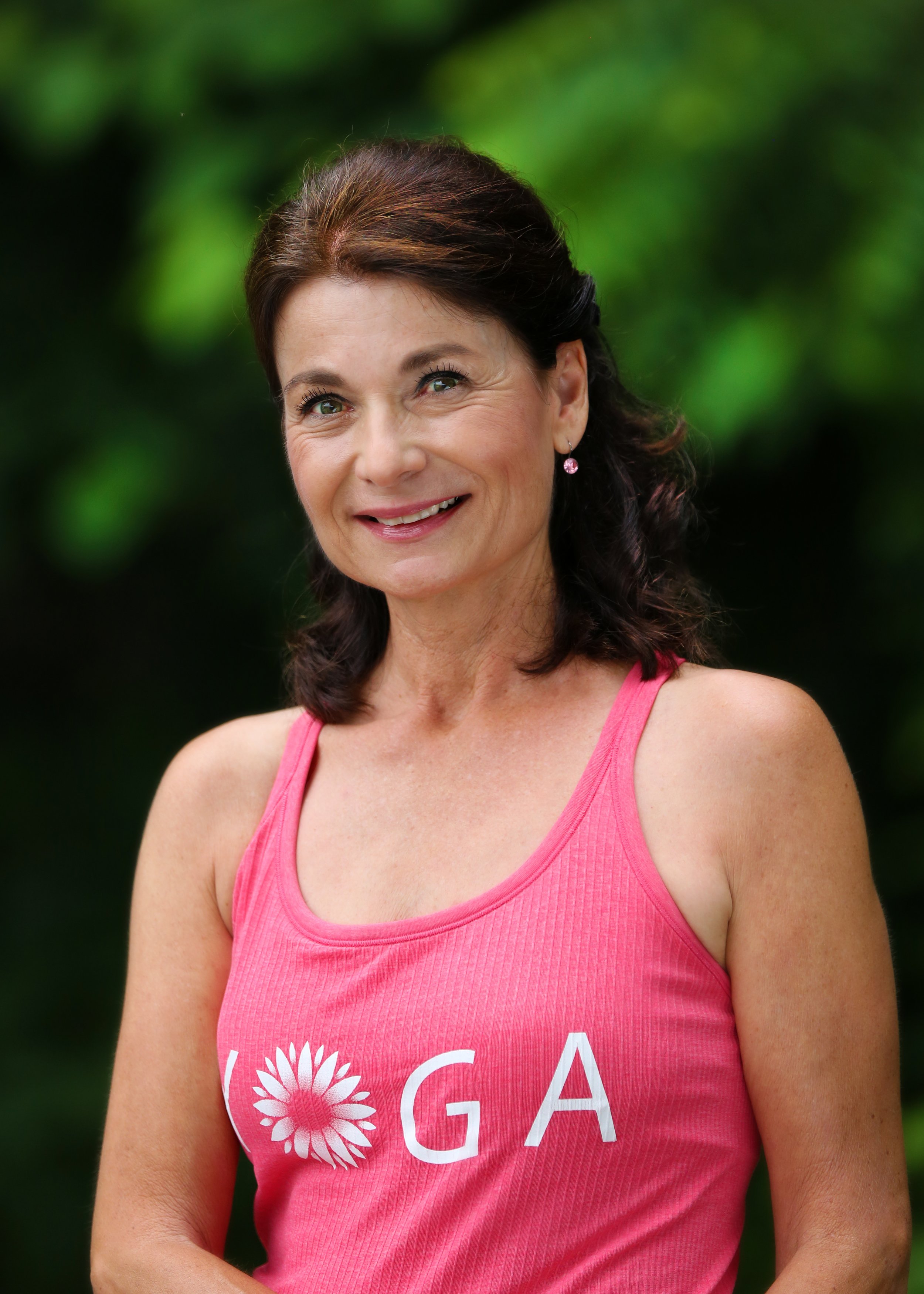 Smiling woman with dark hair in a pink tank top with white text and flower graphic, outdoors against green foliage.