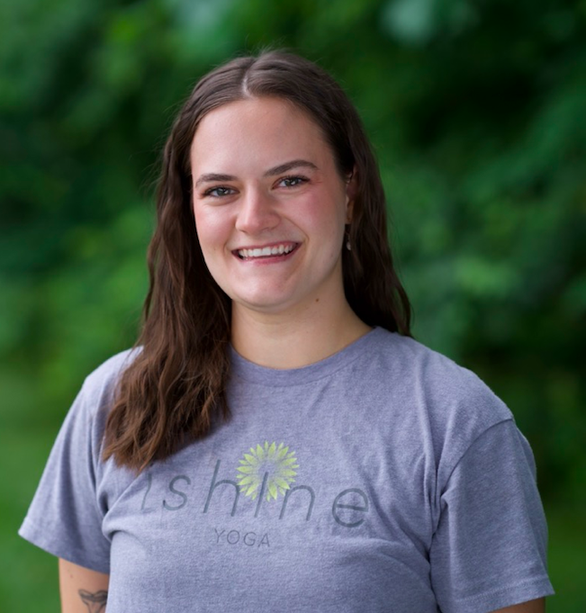 Young woman with brown hair smiling outdoors, wearing a gray t-shirt with a yellow flower design and the word 'shine' on it.
