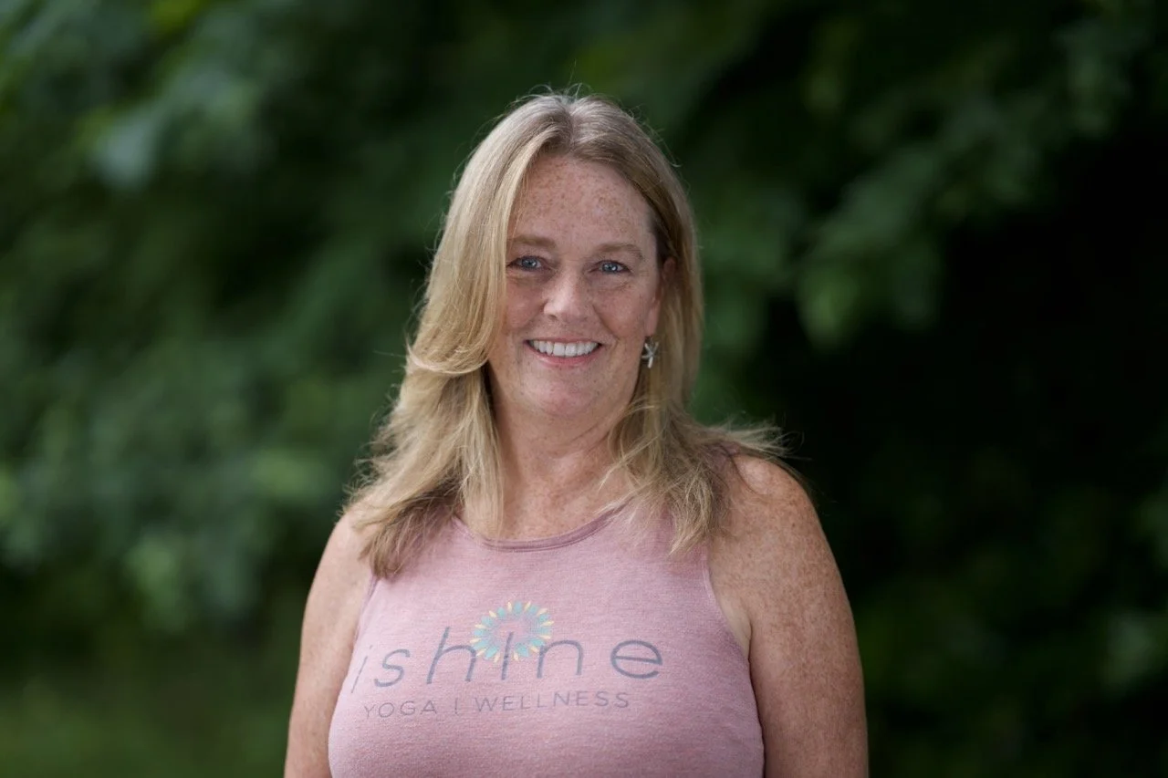 A woman with blonde hair wearing a pink tank top with the words 'shine yoga wellness' on it, standing outdoors with green foliage in the background and smiling at the camera.