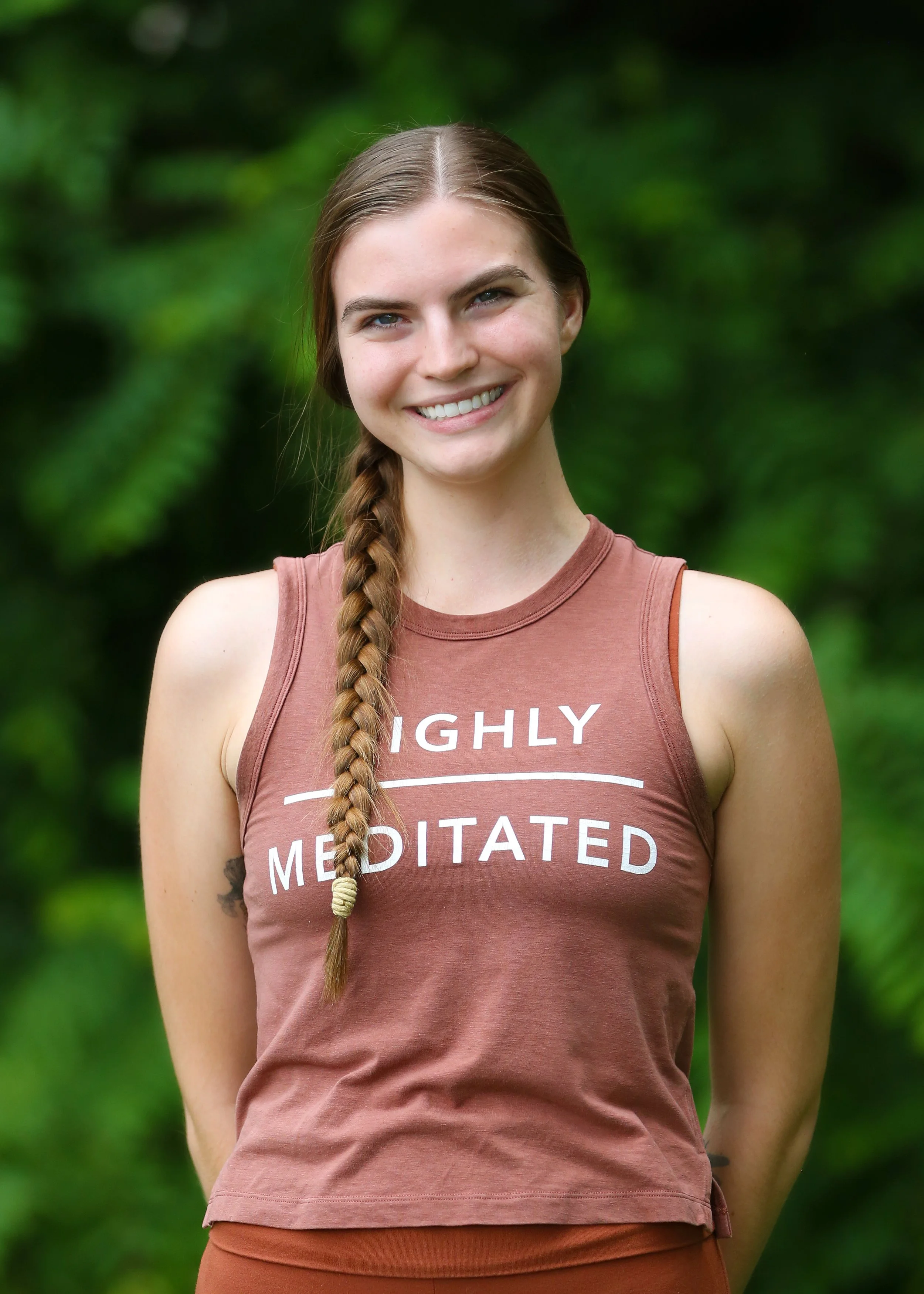 Smiling young woman with long braided brown hair, wearing a sleeveless rust-colored shirt that says 'Highly Meditated', standing outdoors with blurred green foliage in the background.