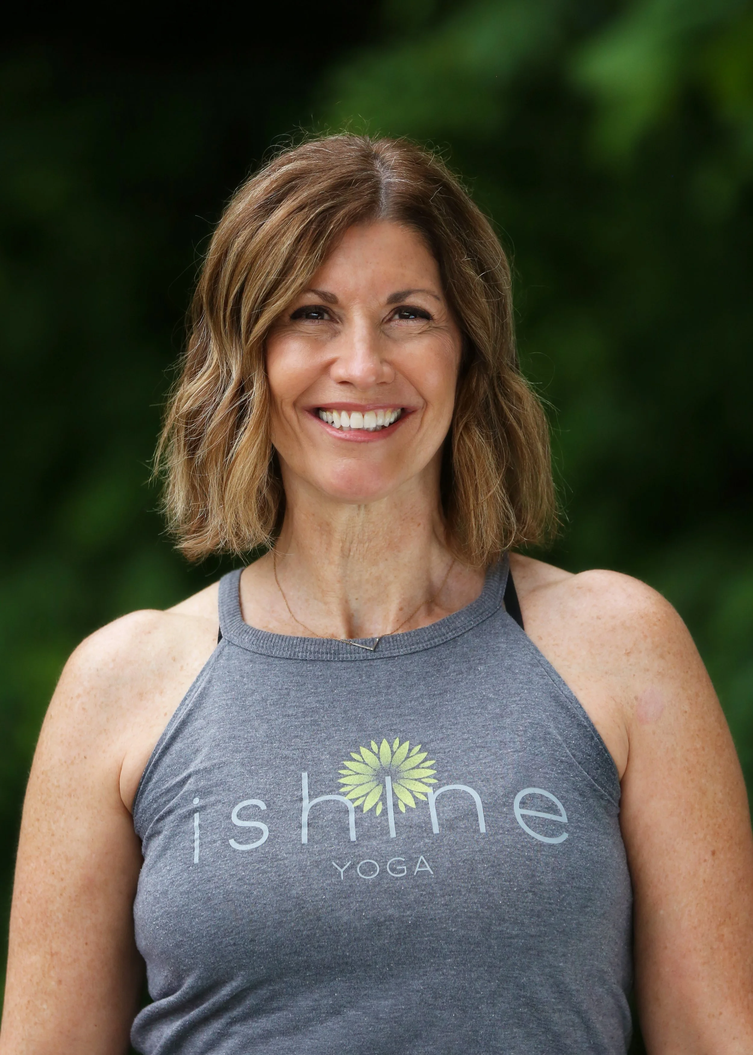 Happy woman with shoulder-length brown hair wearing a gray tank top with 'ishine YOGA' logo, outdoors with green foliage background.