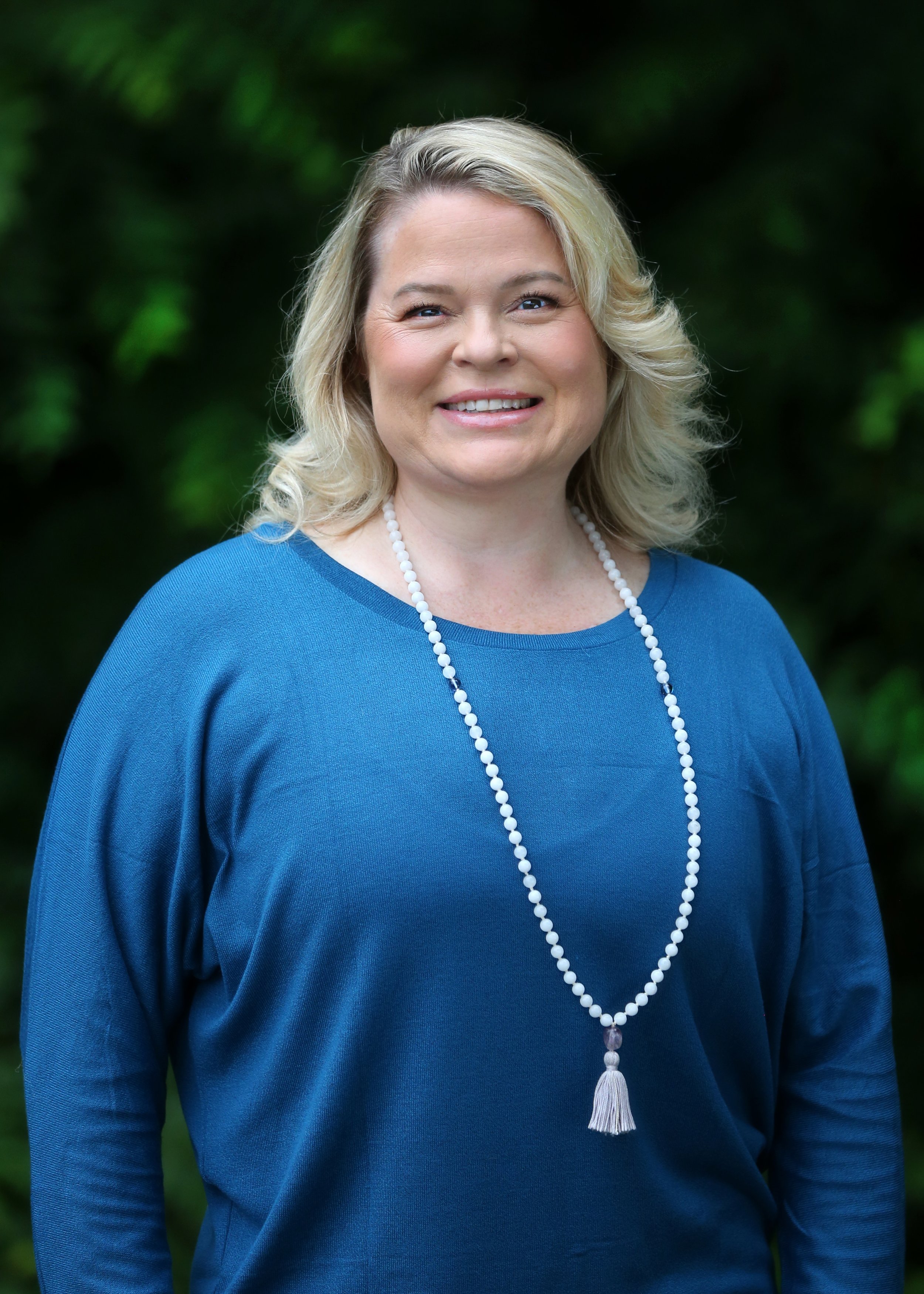 Portrait of a smiling woman with blonde hair, wearing a blue top and a long white beaded necklace with a tassel, standing outdoors with a blurred green leafy background.