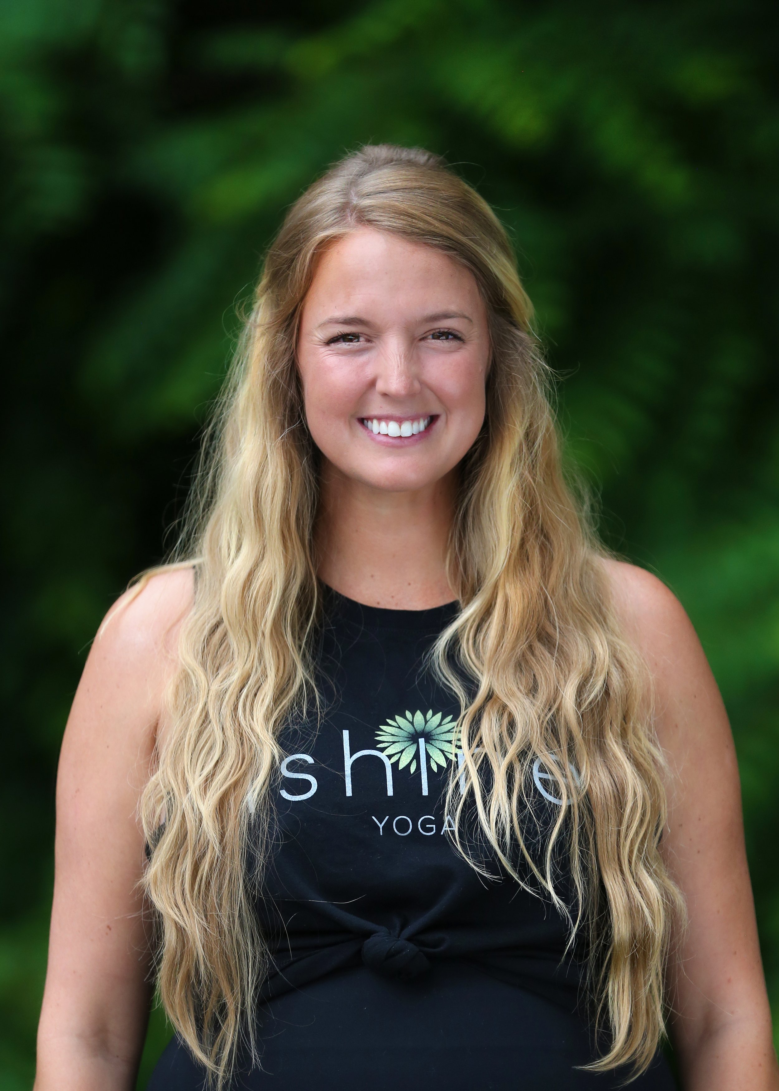 A smiling woman with long wavy blonde hair, wearing a black tank top with a logo that reads 'shift' and 'yoga,' standing outdoors with blurred green foliage in the background.