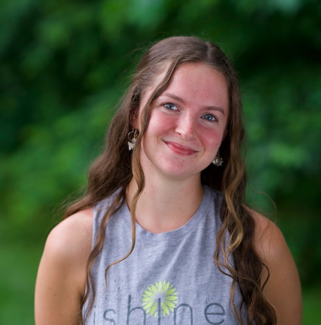A young woman with long brown hair, wearing a grey sleeveless shirt and star-shaped earrings, smiling outdoors with a blurred green background.