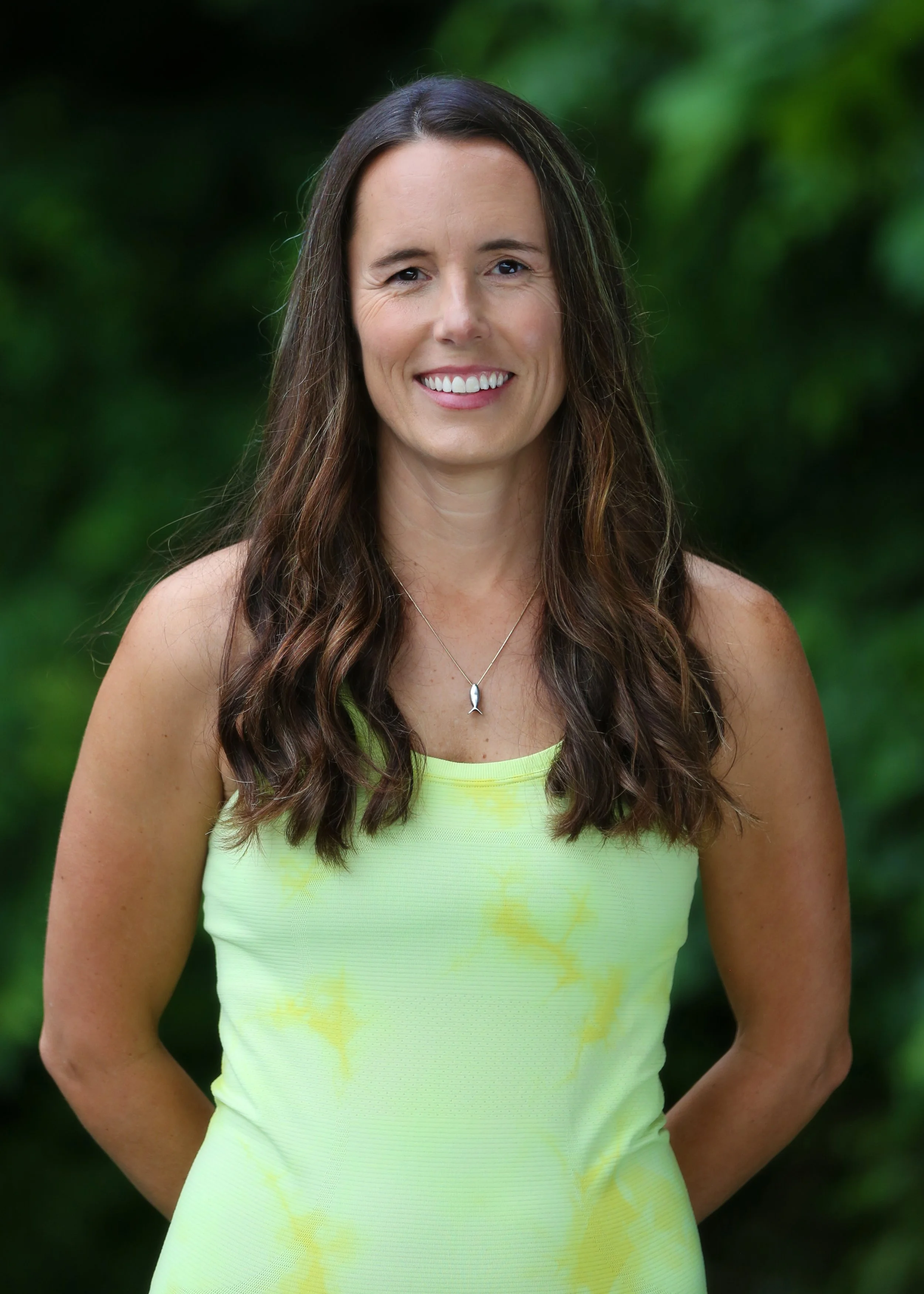 A woman with long brown hair in loose waves, smiling, wearing a bright yellow sleeveless top and a silver necklace with a fish pendant, standing outdoors with green foliage in the background.