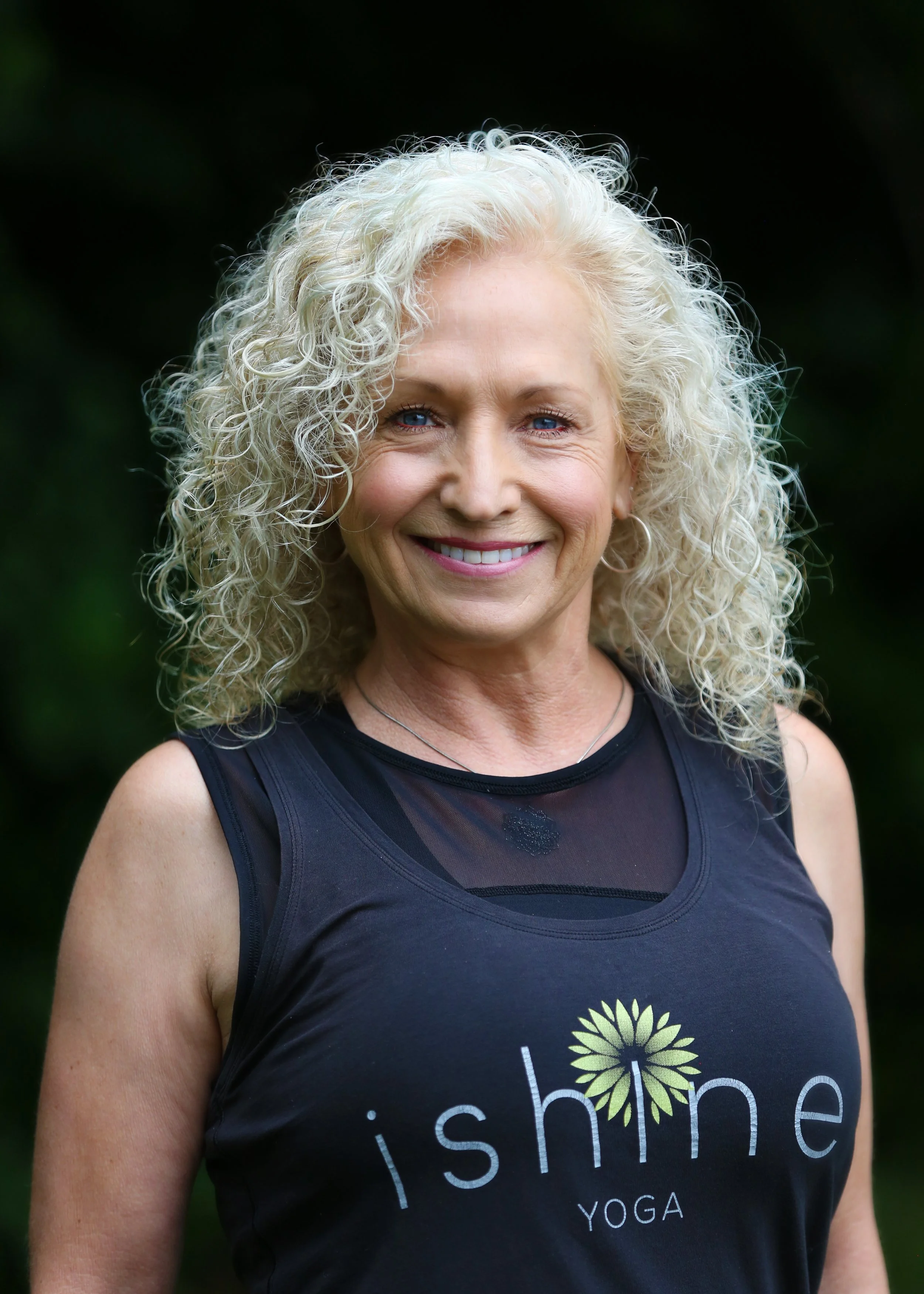 A smiling older woman with curly gray hair, wearing a black sleeveless shirt with the words 'i shine' and a sunflower graphic on it, standing outdoors with a blurred green background.