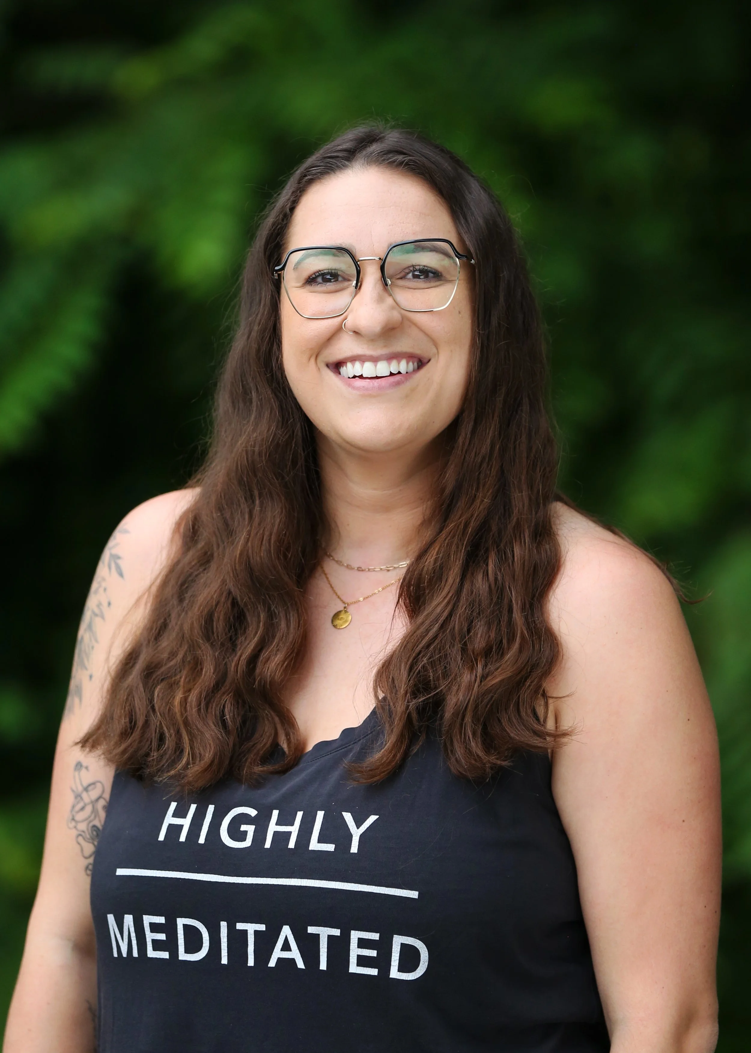 A woman smiling outdoors with green foliage background, wearing glasses, a nose ring, a black tank top that says 'Highly Meditated,' and layered necklaces.
