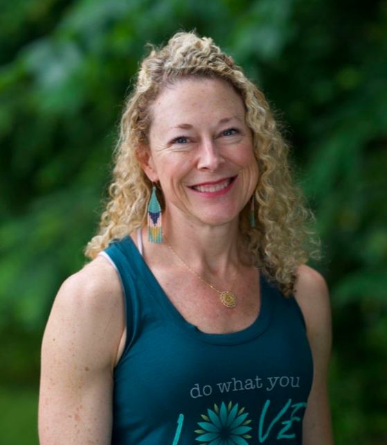A woman with curly blonde hair smiling outdoors in front of green foliage, wearing a blue tank top and colorful earrings.