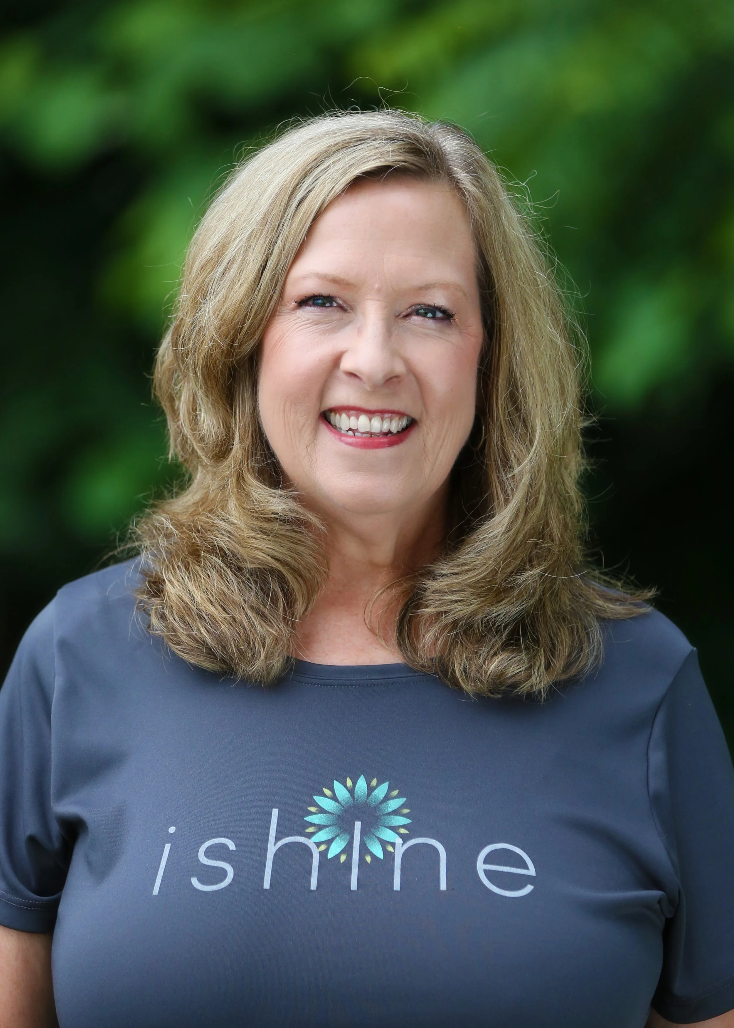Smiling woman with shoulder-length blonde hair wearing a navy T-shirt with the word 'shine' and a flower logo, standing outdoors with green foliage in the background.