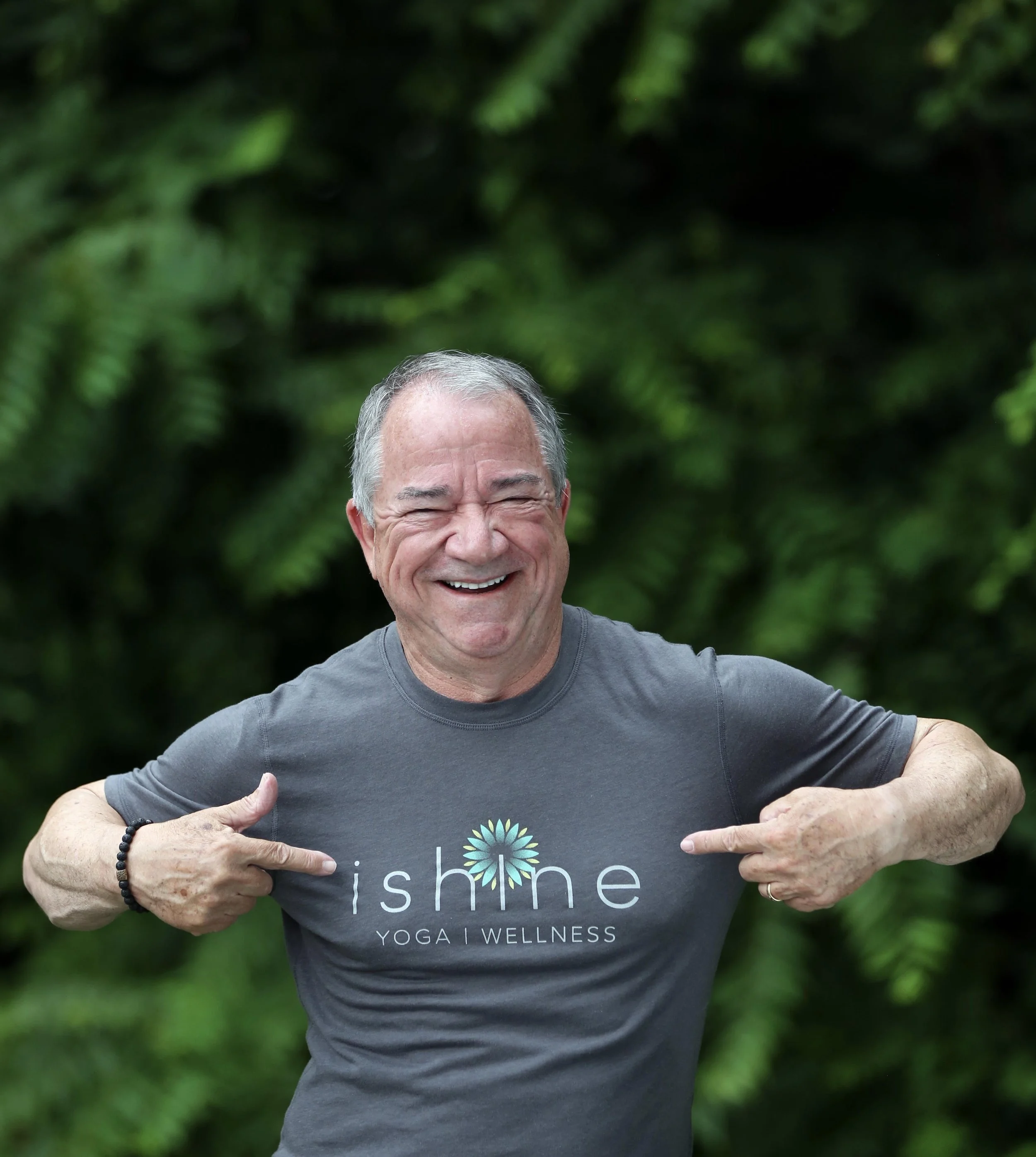 A smiling older man points at his gray t-shirt that has the words 'shine Yoga Wellness' with a colorful, circular logo. He is outdoors with a lush green background.
