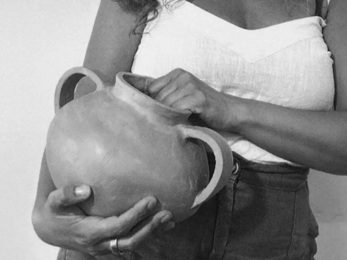 A person holding a ceramic kettle with an open lid, wearing a white top and dark pants.