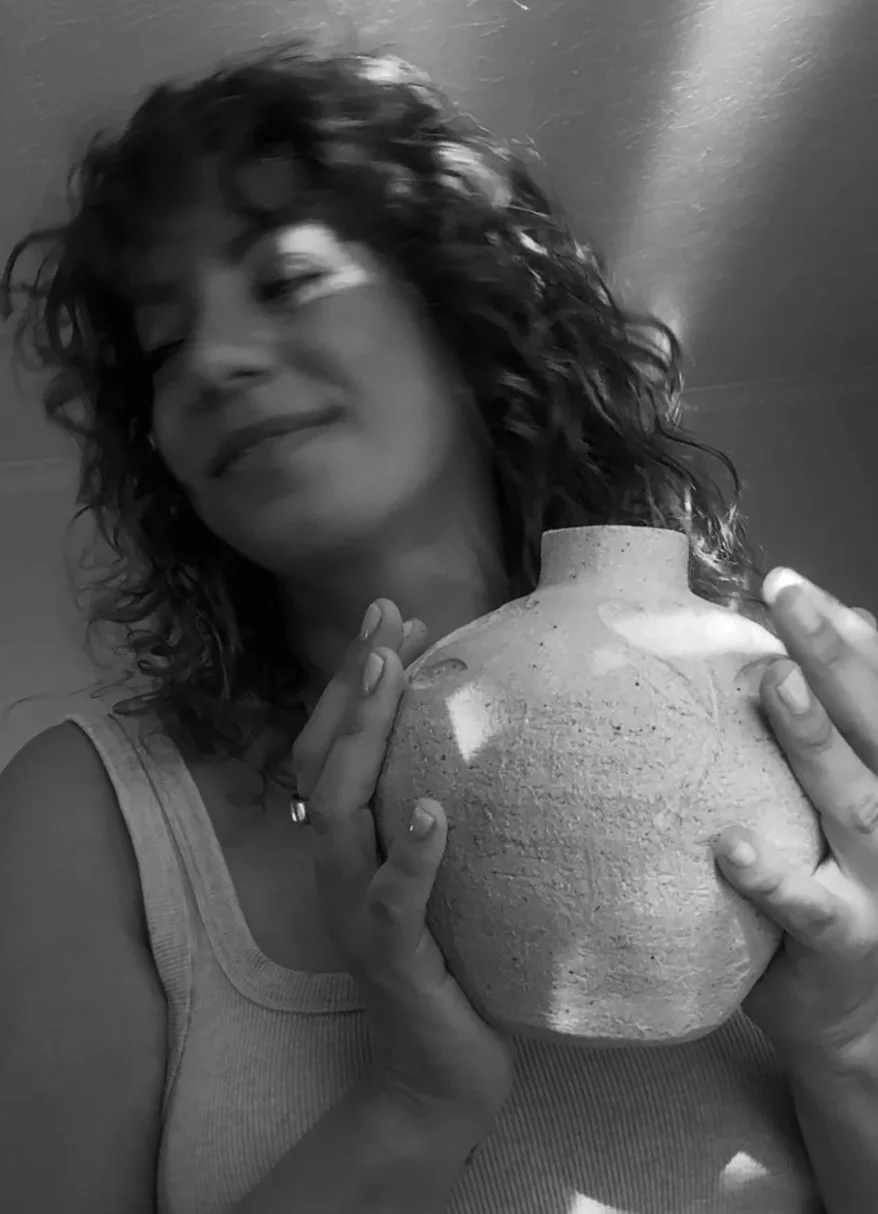A woman with curly hair holding a large ceramic vase, smiling with her eyes closed, in a room with a framed picture on the wall.