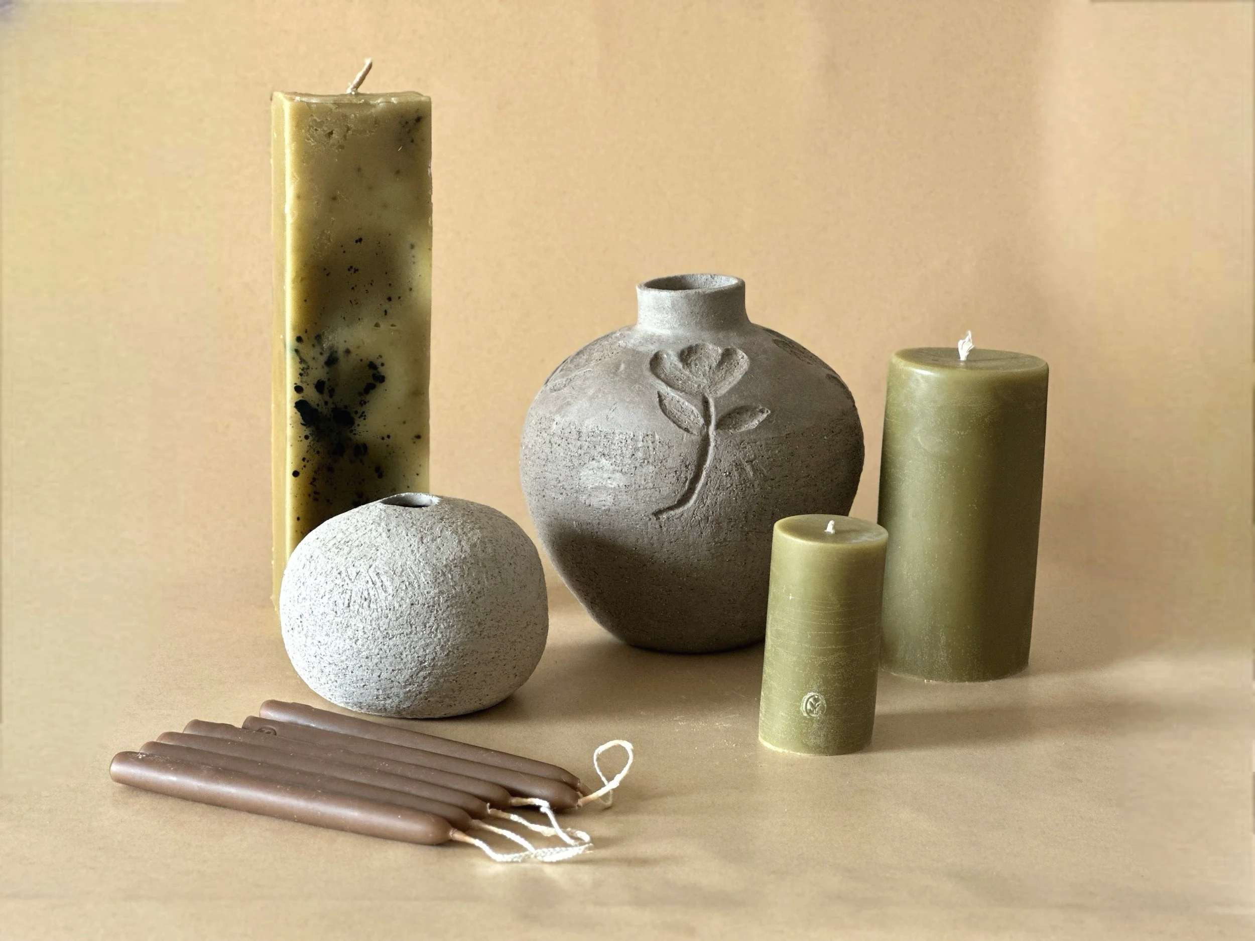 A collection of candles and a ceramic vase on a beige background, including tall yellow, tall green, a textured white sphere, a textured gray sphere with a carved flower, and several thicker beige candles.