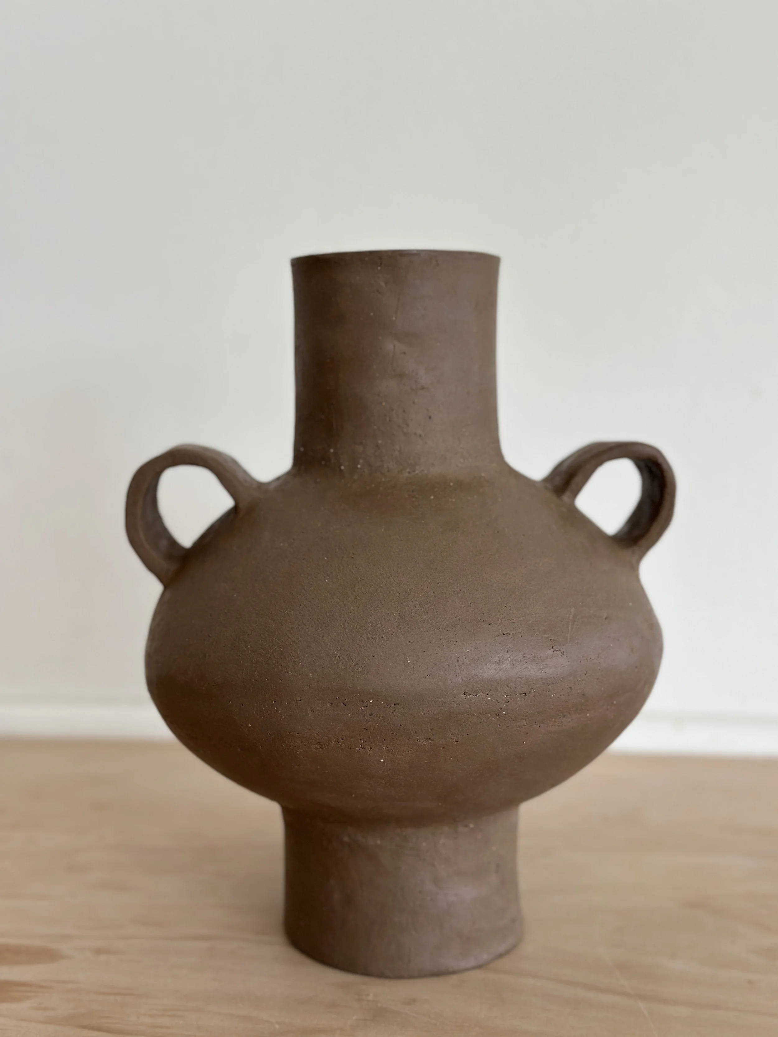 A brown ceramic vase with two small handles, placed on a wooden surface against a plain white background.