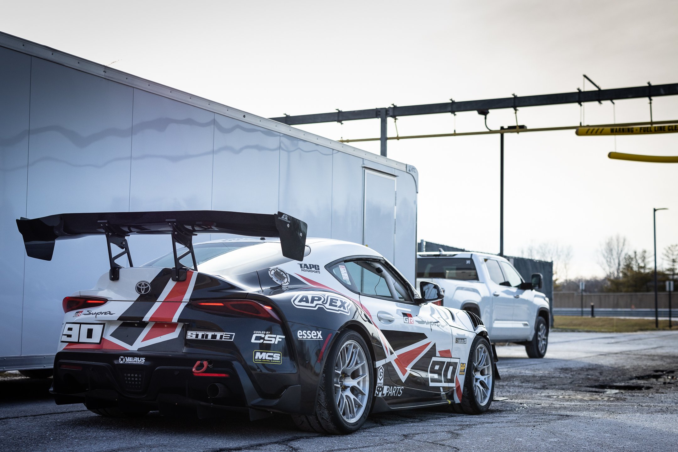 Rear three-quarter view of the number 90 Toyota GR Supra endurance race car parked beside the team trailer and pickup truck