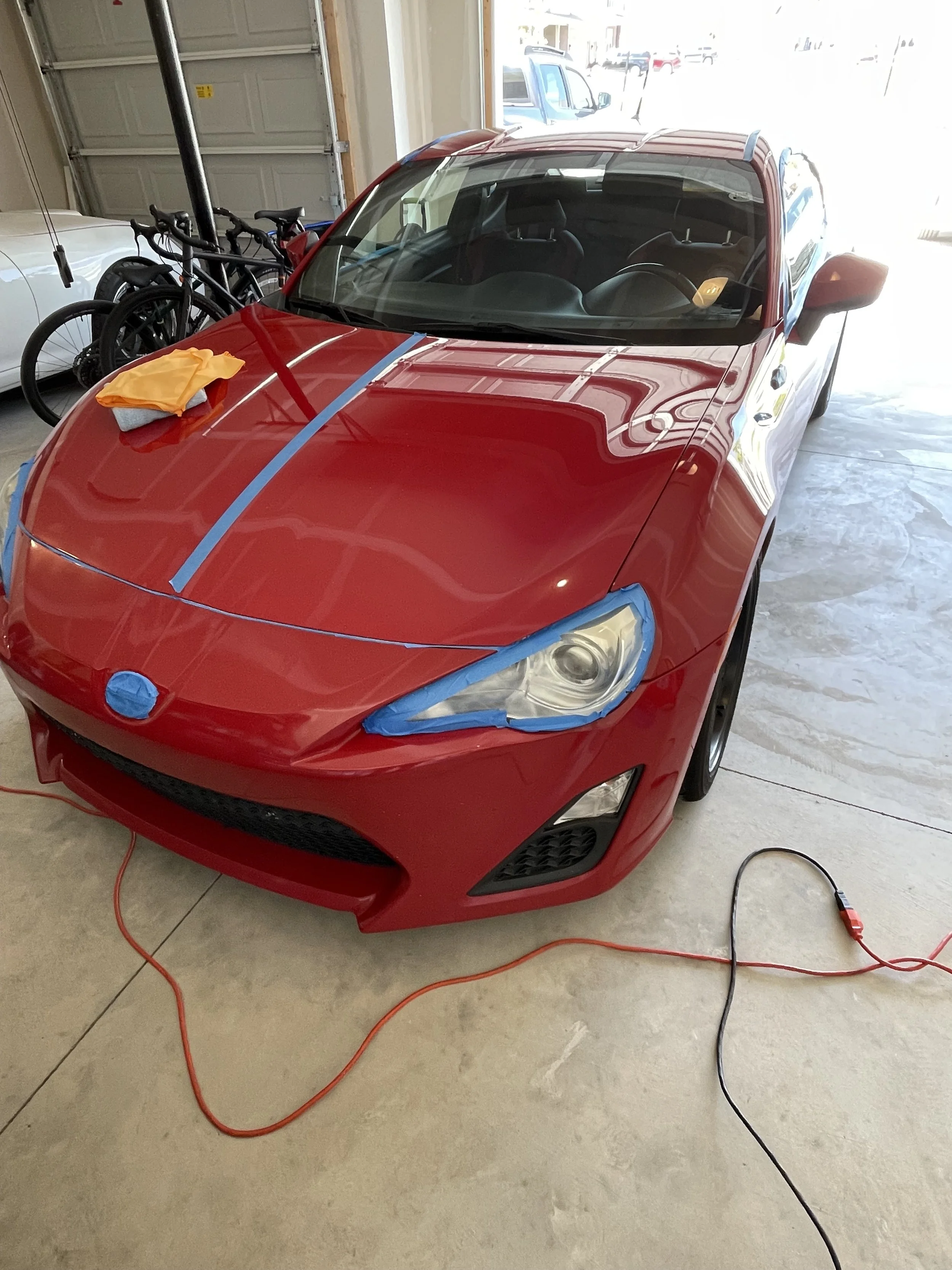 Red Scion FR-S in a garage with blue tape around the headlights and emblem and down the hood for paint correction.