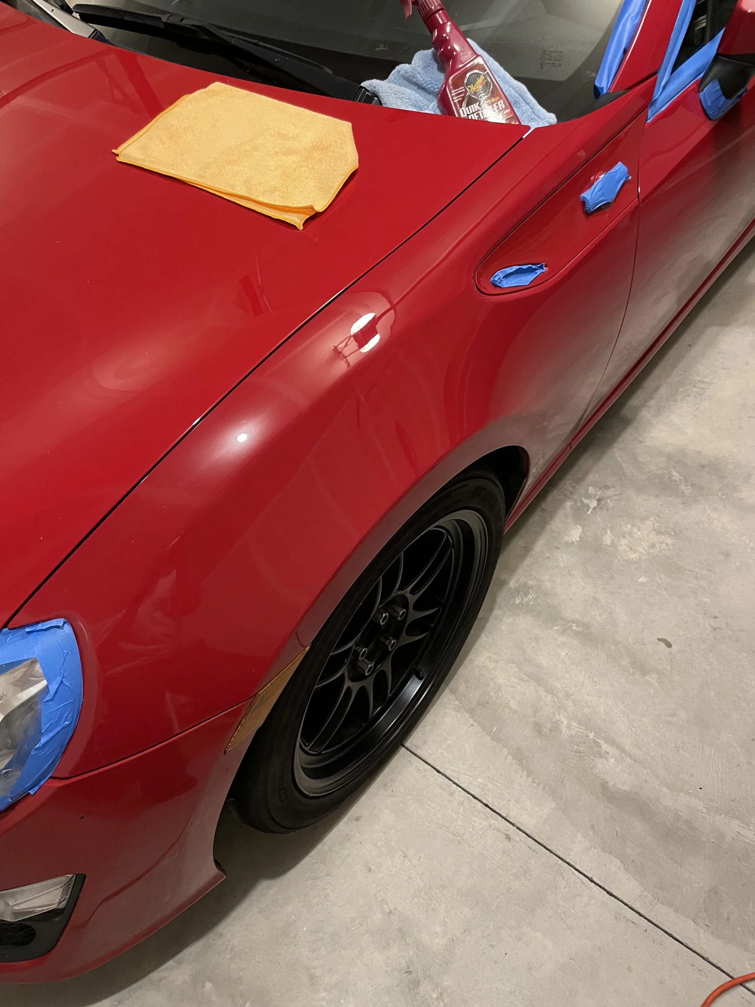 Red FR-S hood and front fender with a yellow microfiber towel and blue masking tape around the headlight.