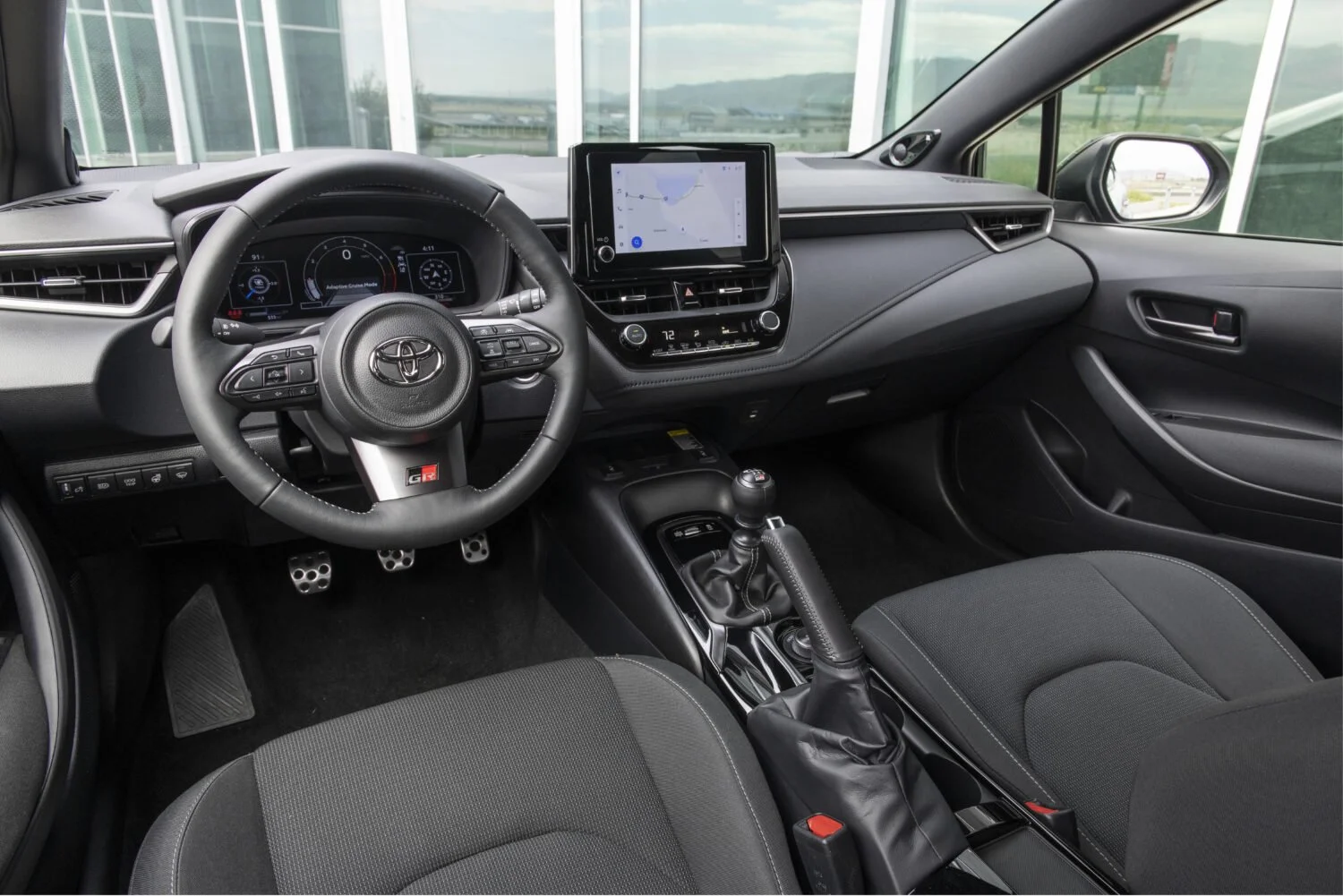 Toyota GR Corolla interior with black front seats, GR steering wheel, digital gauges, center touchscreen, manual gear lever, and handbrake.