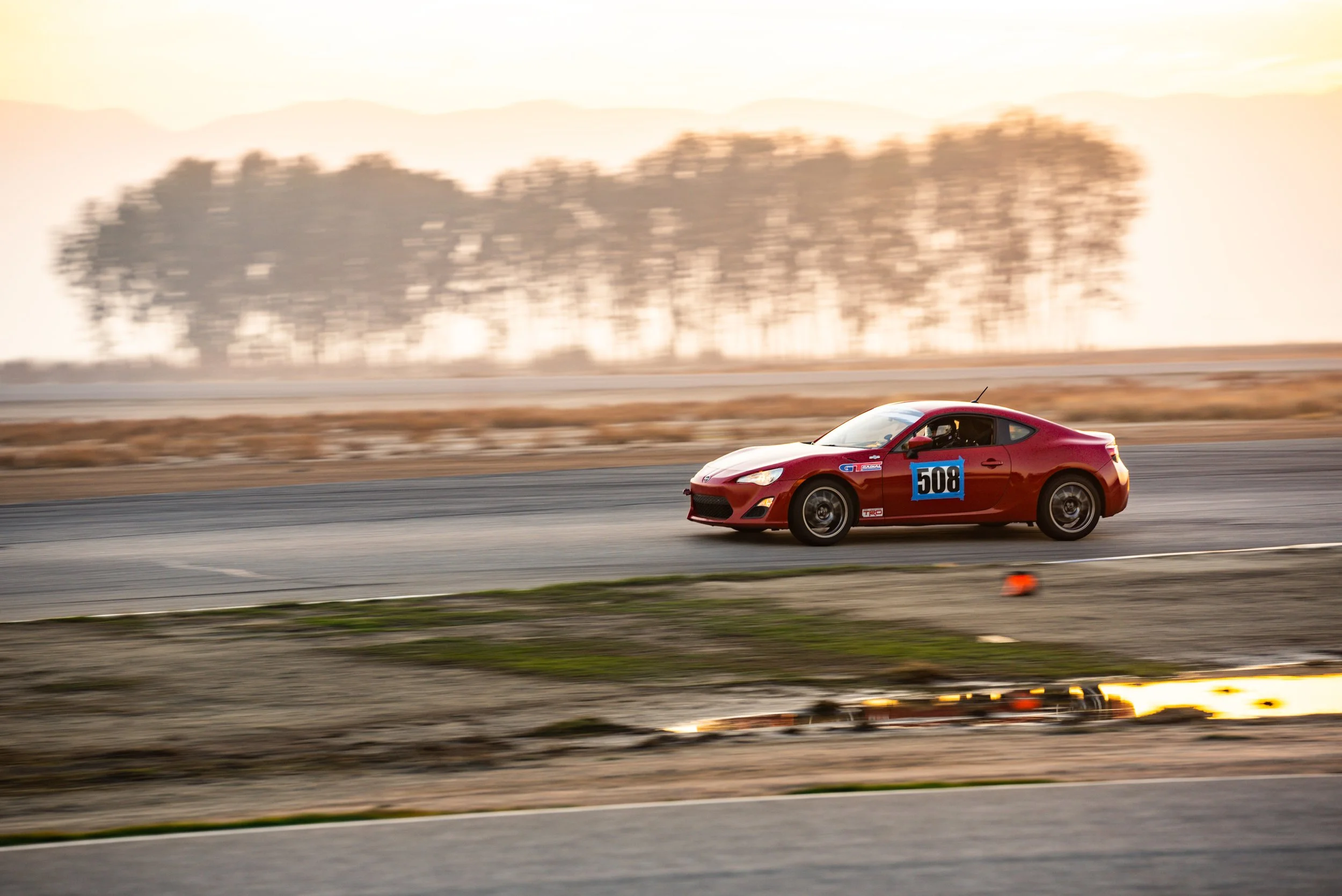 Red Scion FR-S number 508 driving through a corner at Buttonwillow Raceway in warm evening light, with trees and hills in the background.