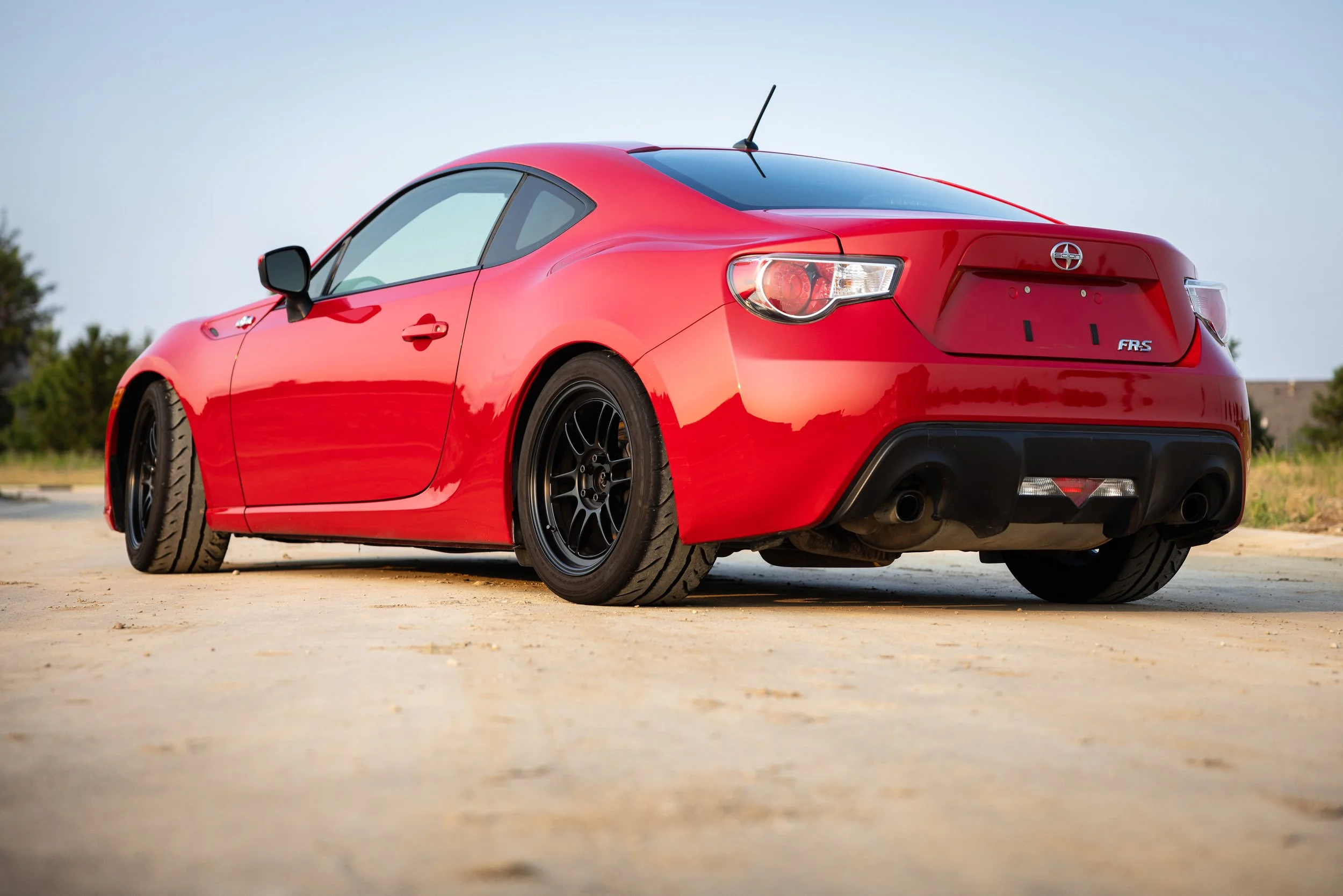 Rear three-quarter view of a red 2013 Scion FR-S with black multi-spoke wheels, parked outdoors on pale concrete.
