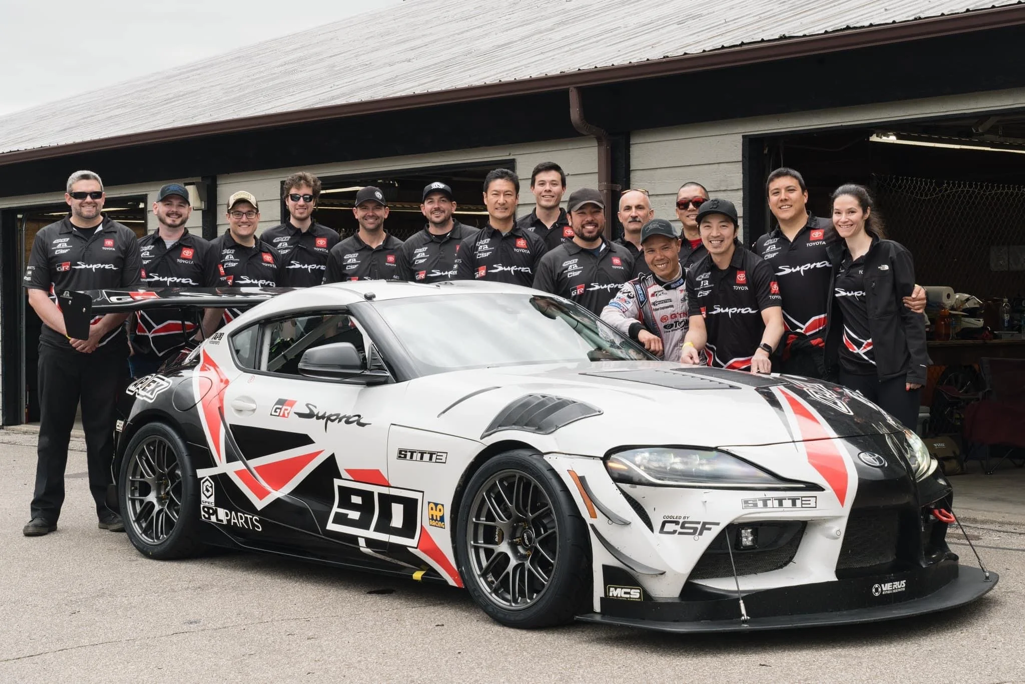 Toyota R&D Racing team gathered behind the number 90 A90 Supra outside the garages at Mid-Ohio Sports Car Course.