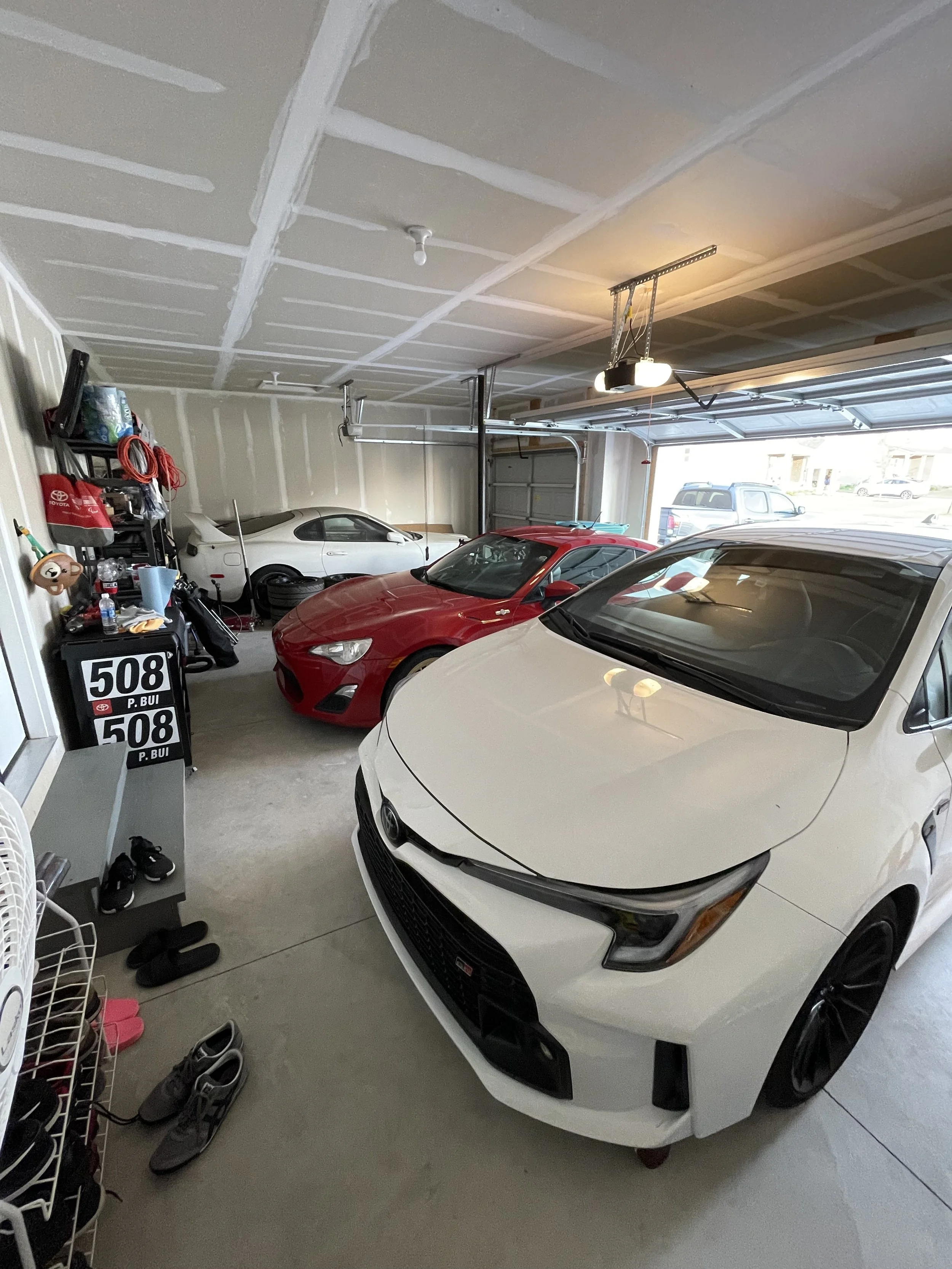 White Toyota GR Corolla at home beside the red Scion FR-S and White MKIV Toyota Supra in the garage and a gray Toyota truck in the driveway.