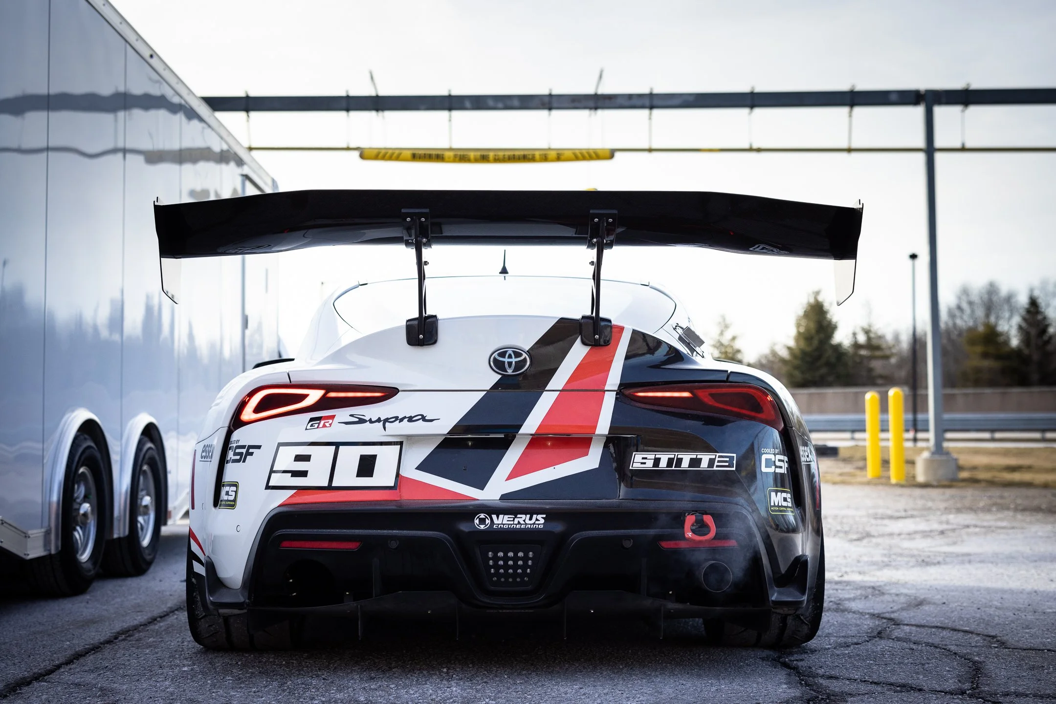Rear view of the number 90 Toyota GR Supra endurance race car with its large rear wing beside the team trailer