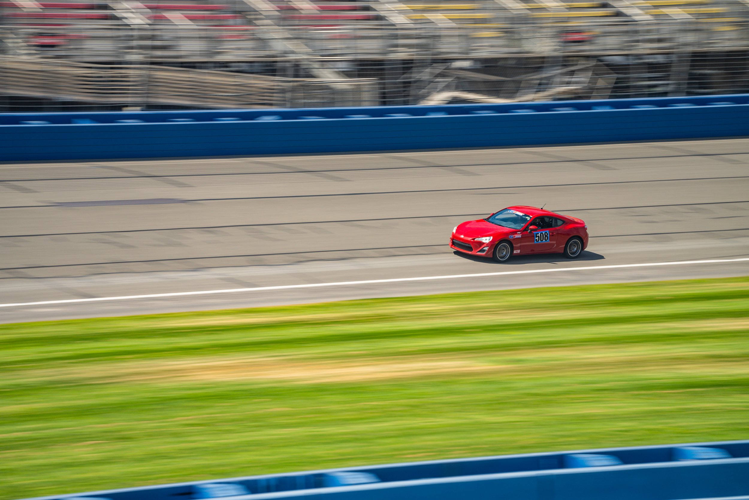 Red Scion FR-S driving along Autoclub Speedway straight beside blue barriers and grandstands, with motion-blurred grass in the foreground.