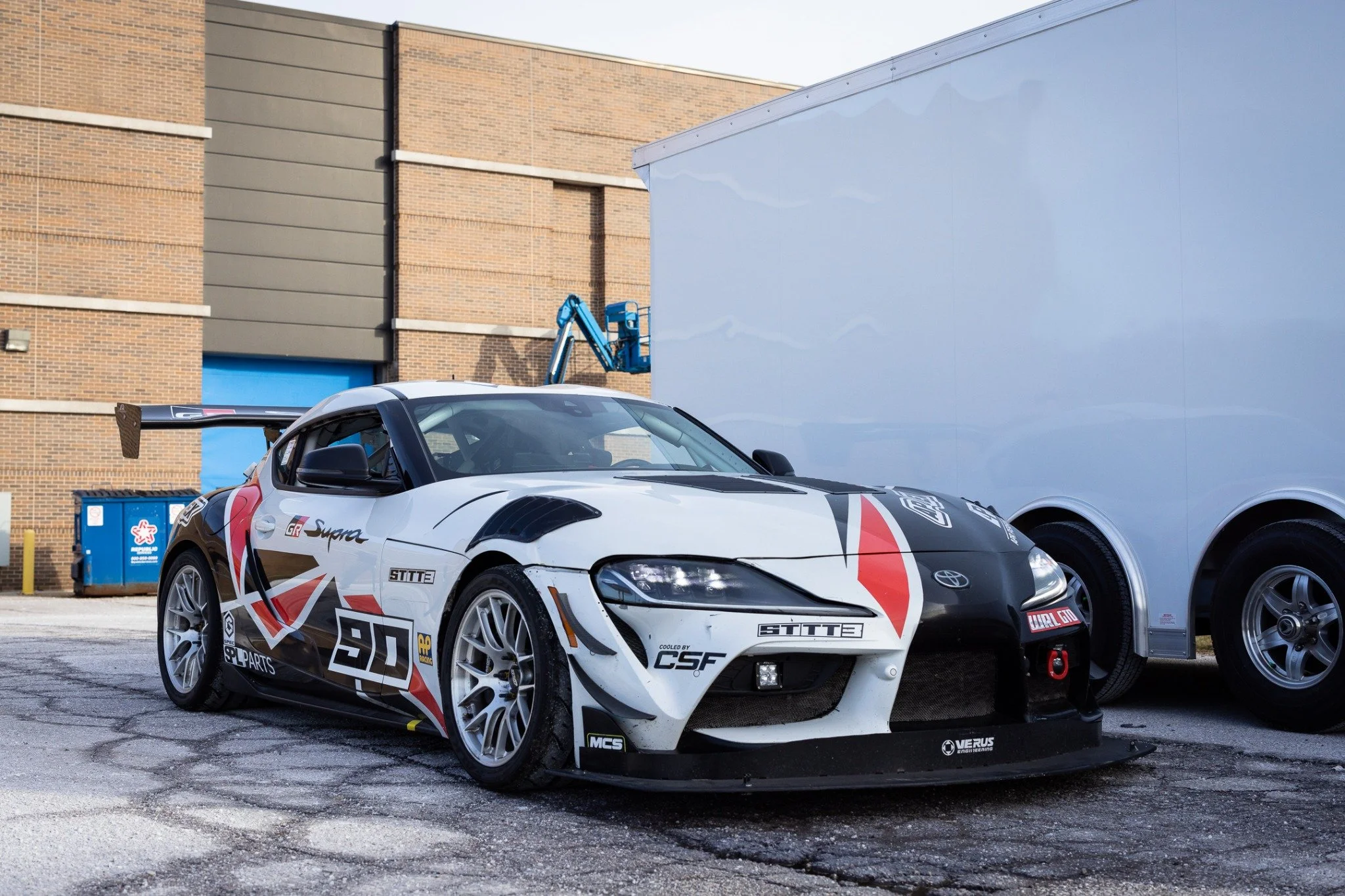 Front three-quarter view of the number 90 A90 Toyota GR Supra endurance race car beside its trailer at Toyota R&D in Michigan