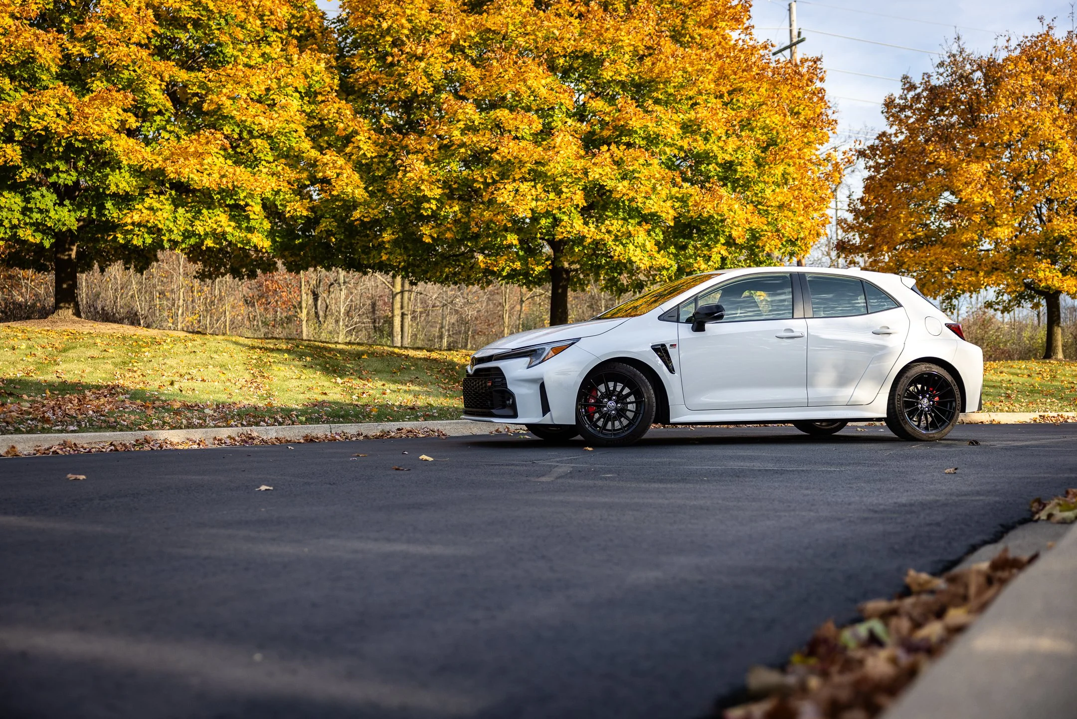 White Toyota GR Corolla with black wheels parked beside a curb beneath yellow and orange autumn trees.