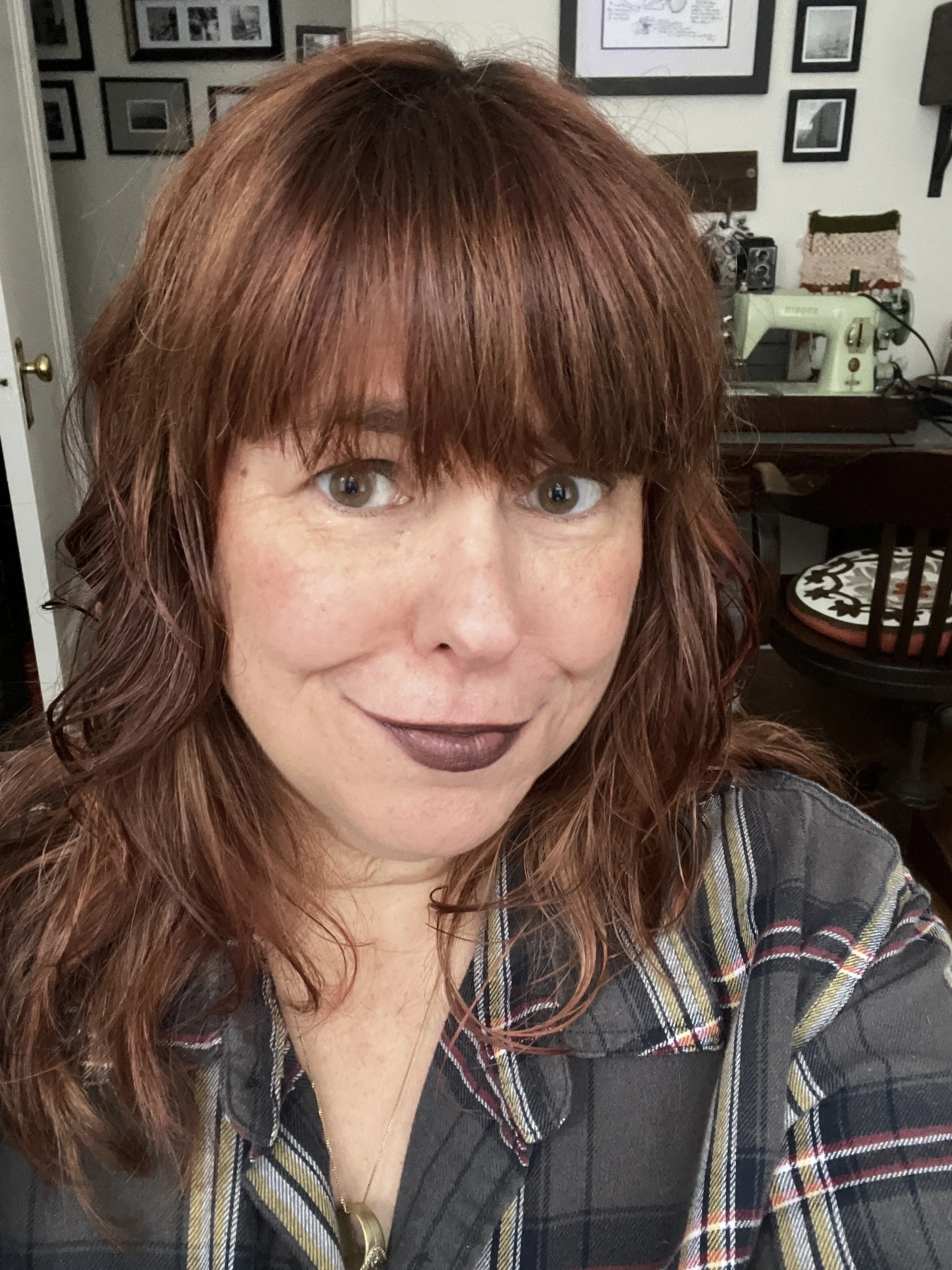 A red haired woman looks smiling into the camera wearing a flannel shirt. In the background on a desk is a sewing machine.