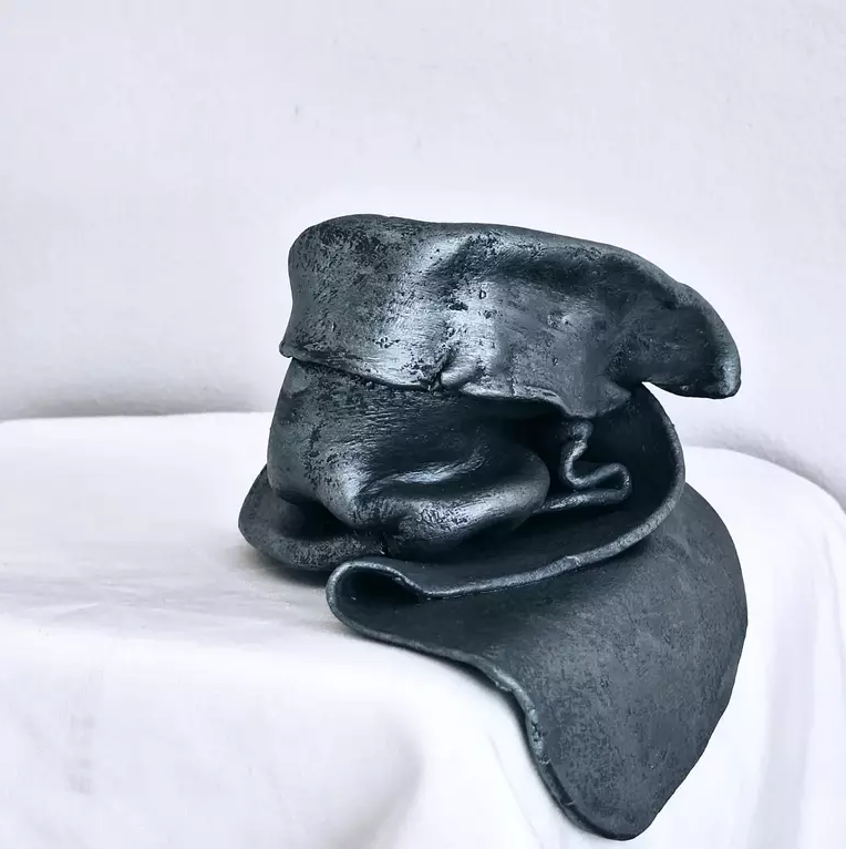 A black abstract sculpture resembling a curled figure resting on a white cloth surface against a plain wall background.