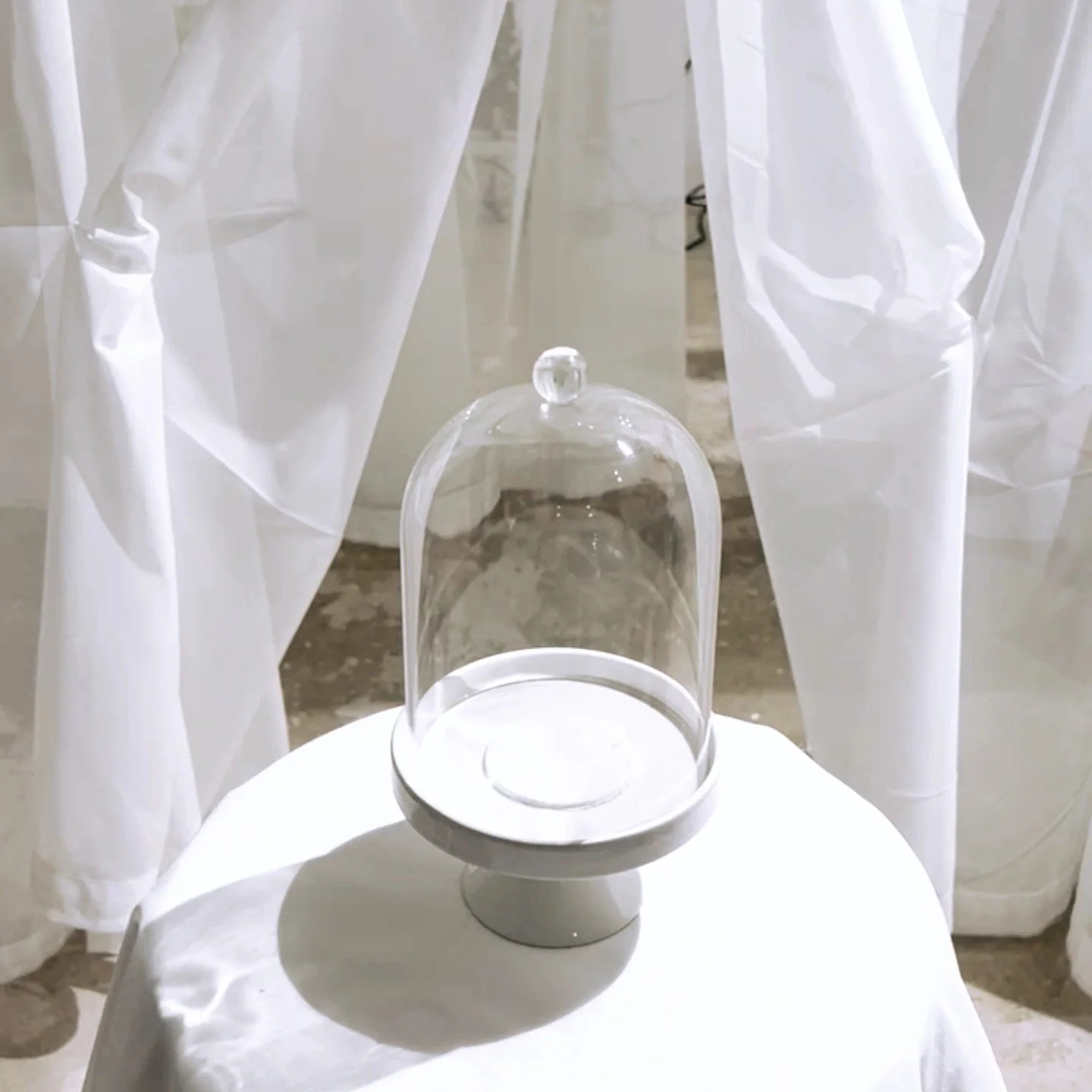A glass dome on a white pedestal, placed on a small white table, with sheer white curtains in the background.